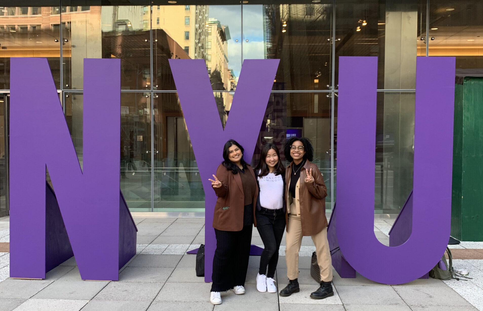 Three female-presenting students of color stand in front of life-size letters spelling “NYU.”