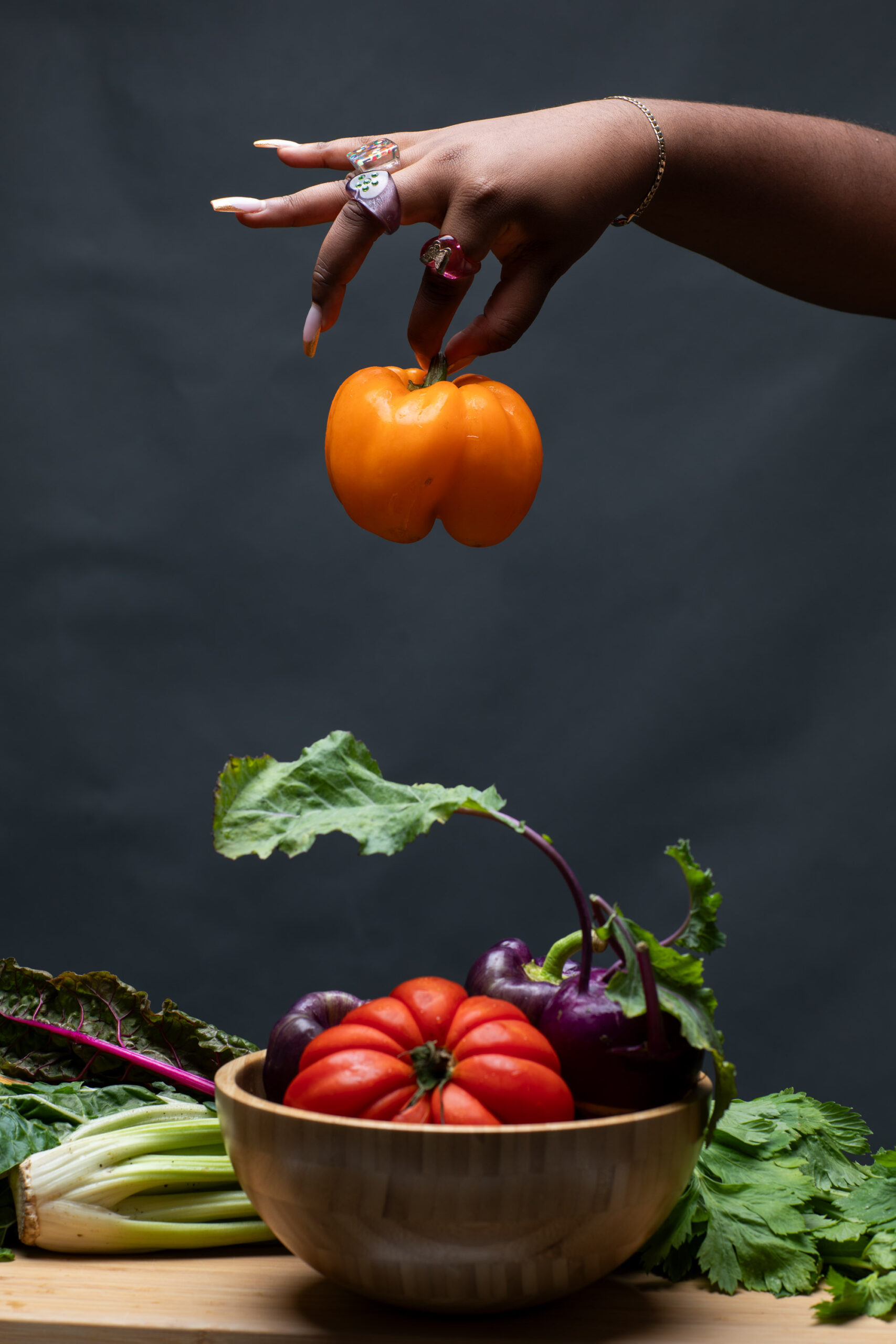 A hand holding an orange bell pepper over a bowl of multicolored vegetables.
