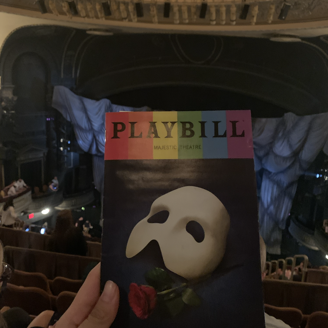Hand holding a Phantom of the Opera playbill in a theatre auditorium.