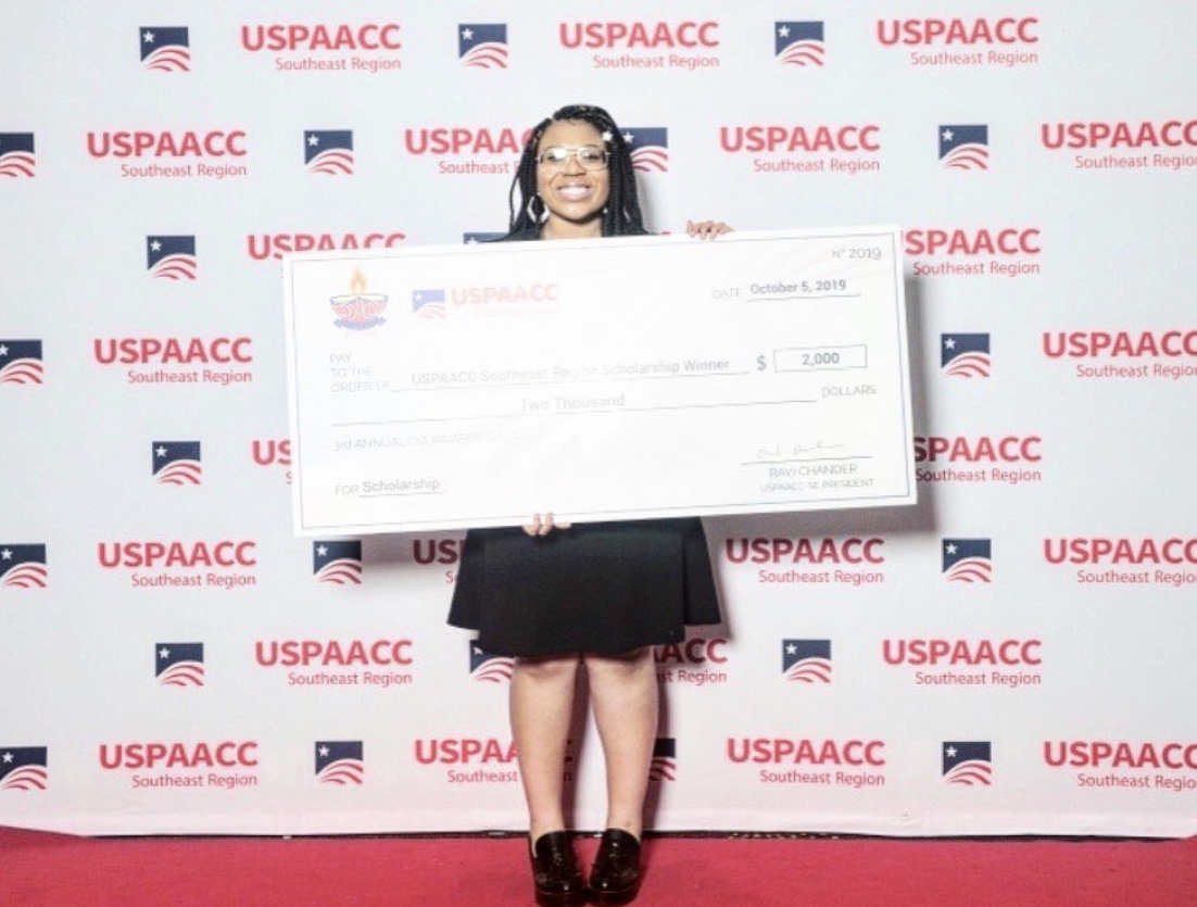 A Black woman in a black dress holds up a scholarship check.