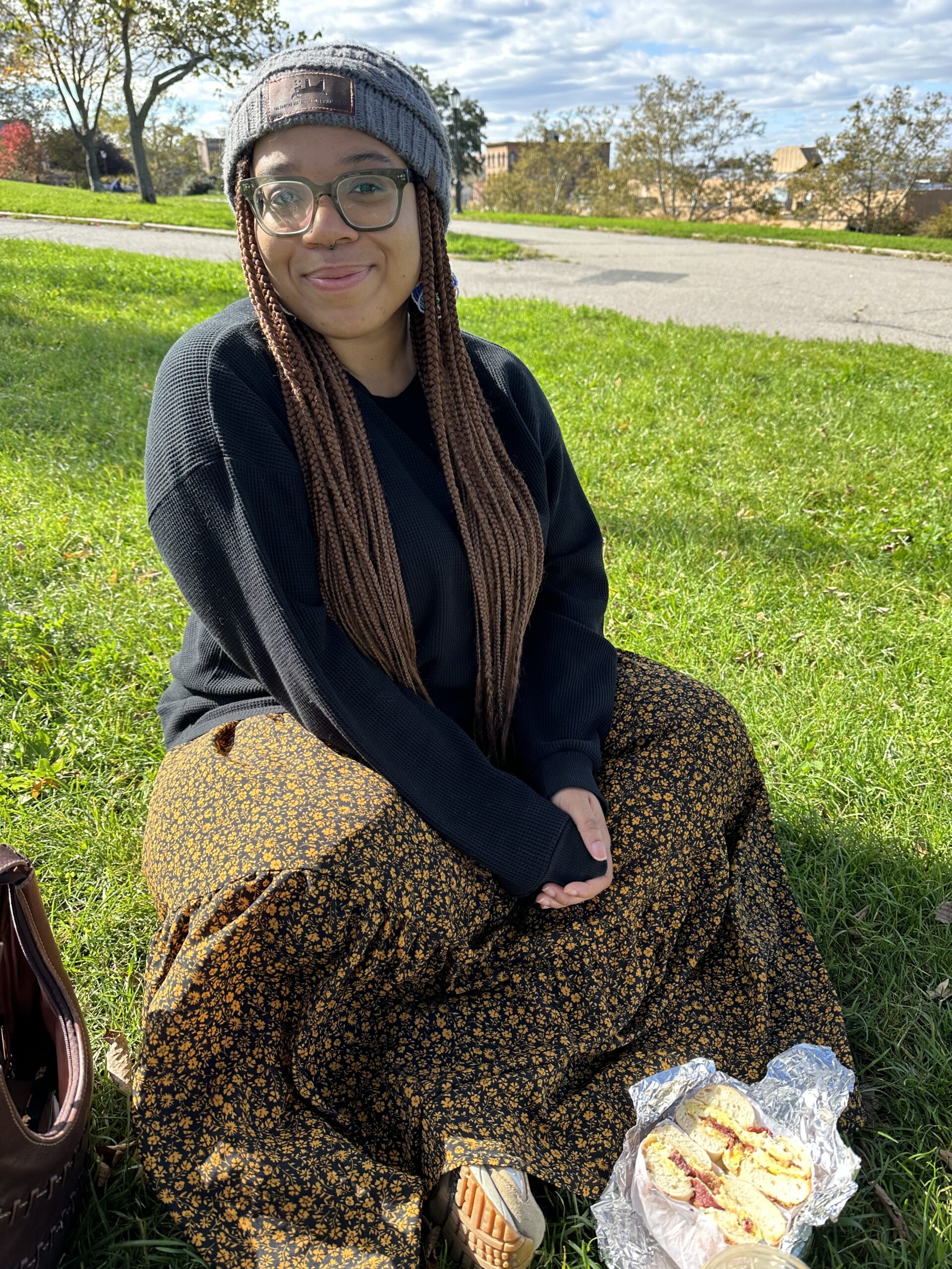 A Black person in a gray beanie, black long sleeve, and orange and black dress sits cross-legged in a park.