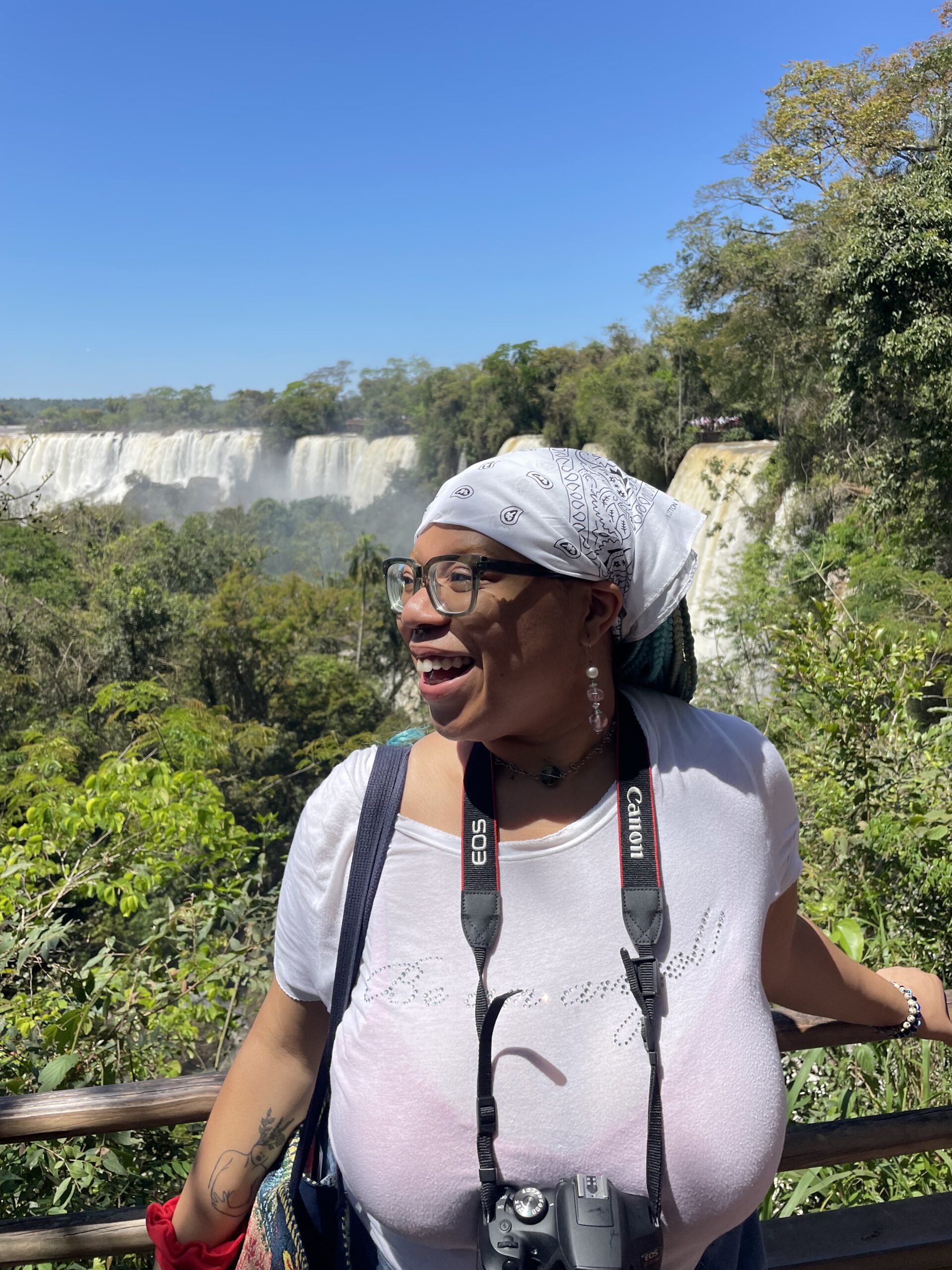 A Black person in a white top and white bandana turns to the side and smiles with greenery and a waterfall flowing behind them in Buenos Aires.