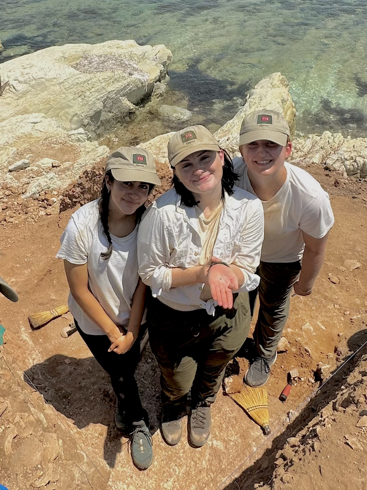Three female-presenting students stand on an archeological dig site where they are conducting research. The person in the middle holds an artifacts in their hand.