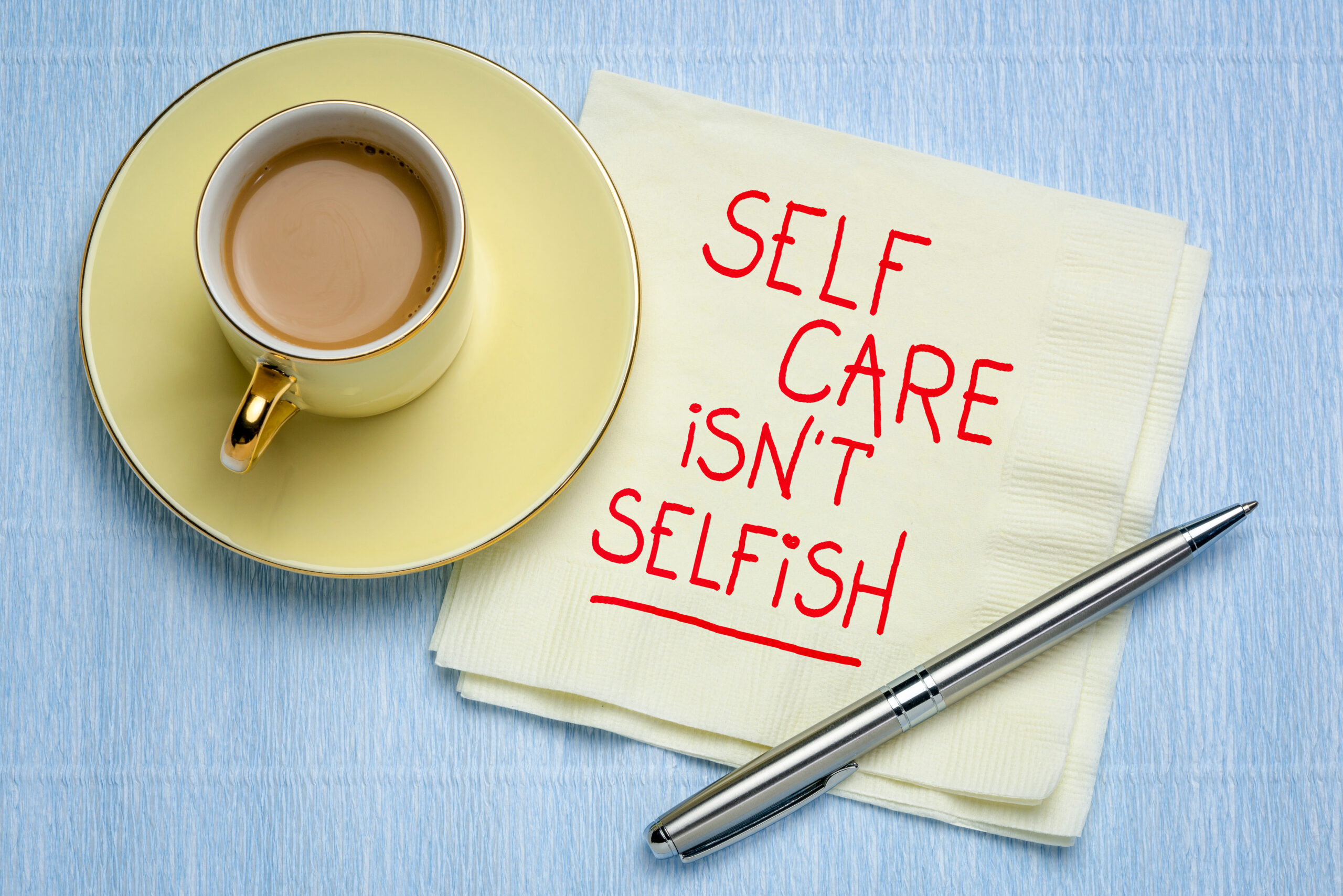 Red handwriting on a napkin that says, “Self care isn’t selfish,” with a cup of coffee on the side.
