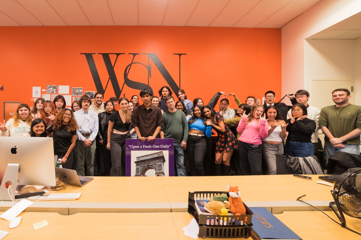 A group of students stand in front of an orange wall decorated with the painted letters “WSN.” WSN (Washington Square News) is NYU’s independent student newspaper.