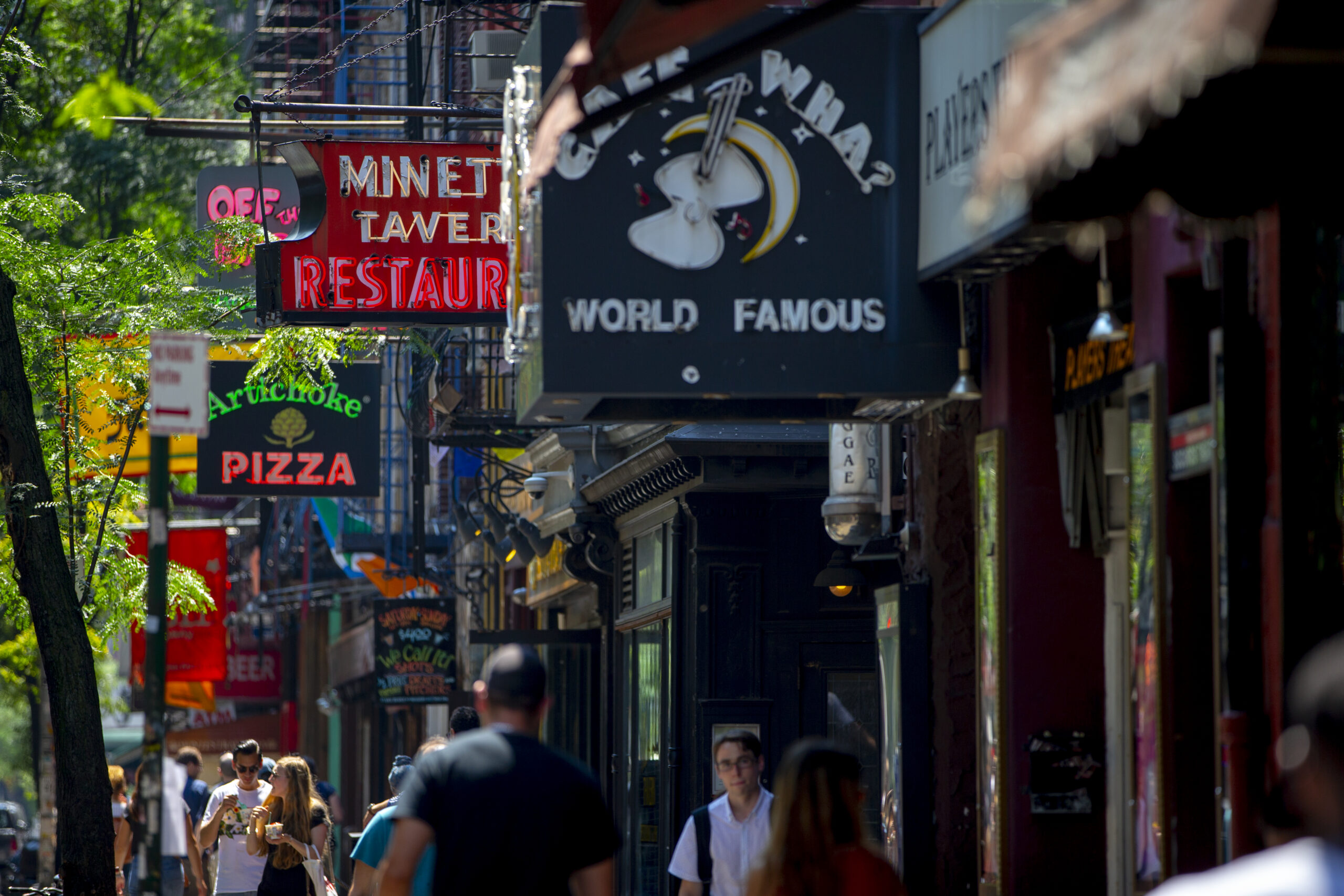 Busy Greenwich Village street lined with storefronts featuring vibrant signs elevated above eye level.
