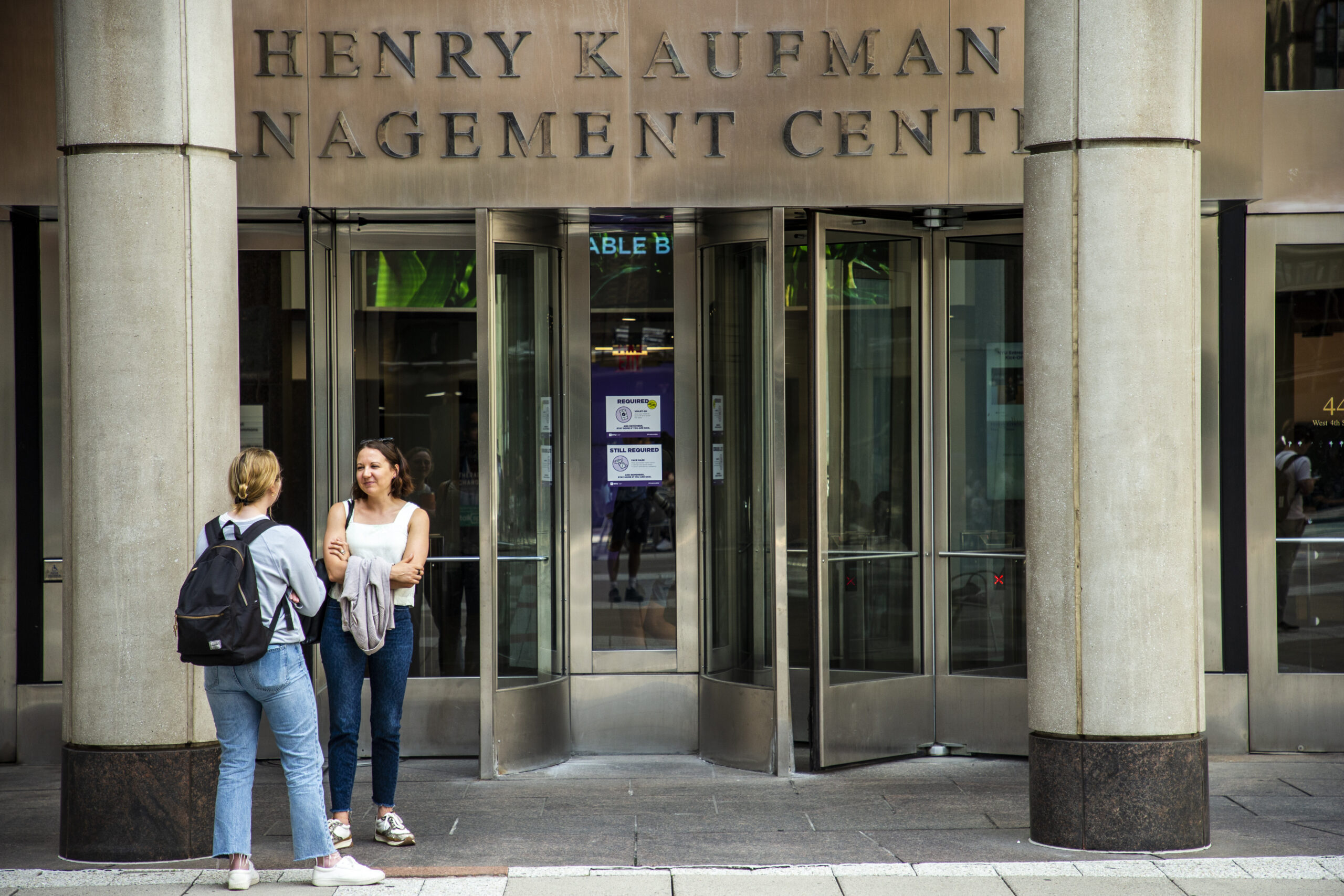Two female-presenting students standing outside the entrance of NYU’s Stern School of Business, the Henry Kaufman Management Center.
