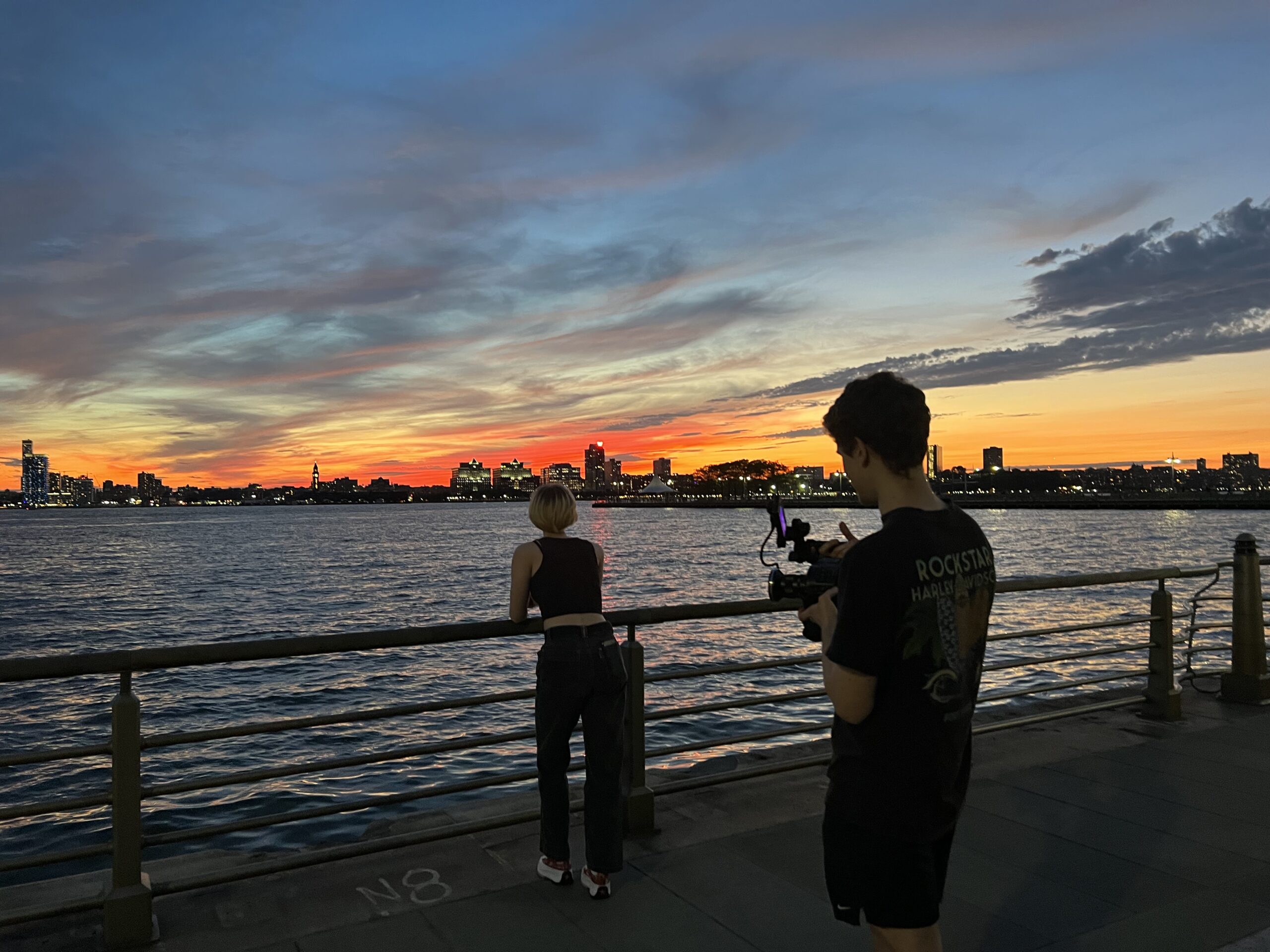 Behind the scenes of the Sight and Sound: Filmmaking production course. Two students stand near a body of water. One is in front of the camera, acting, the other is behind the camera, shooting the scene.