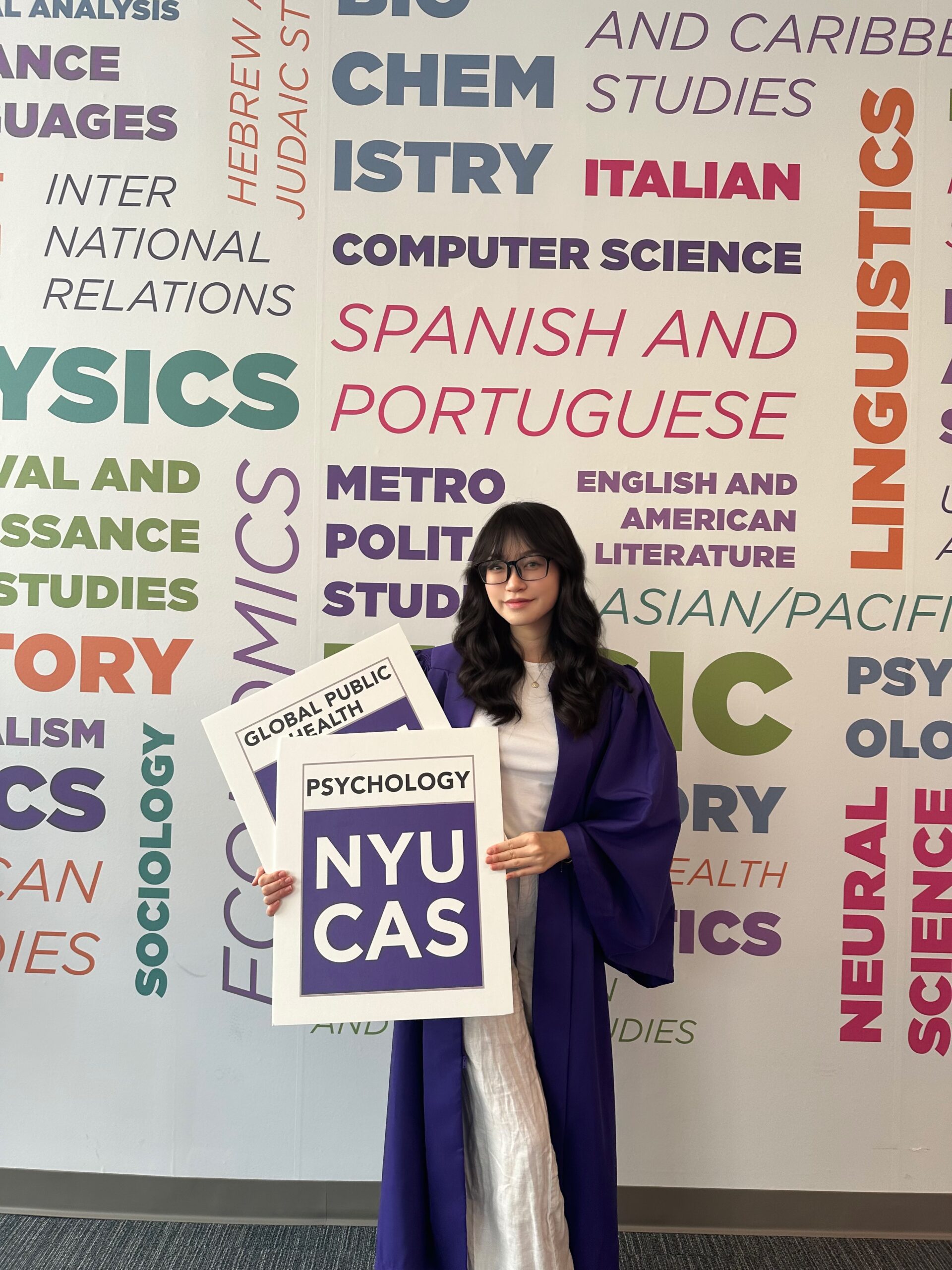 Yasmin Hung, adorned in her graduation attire, holds a sign declaring her affiliation with NYU’s College of Arts and Science, majoring in Psychology.