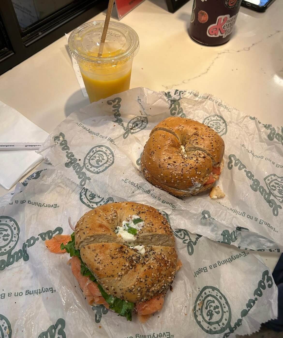 Two toasted everything bagels with plain cream cheese, nova lox, lettuce, red onion, and capers and a cup of orange juice on a table.