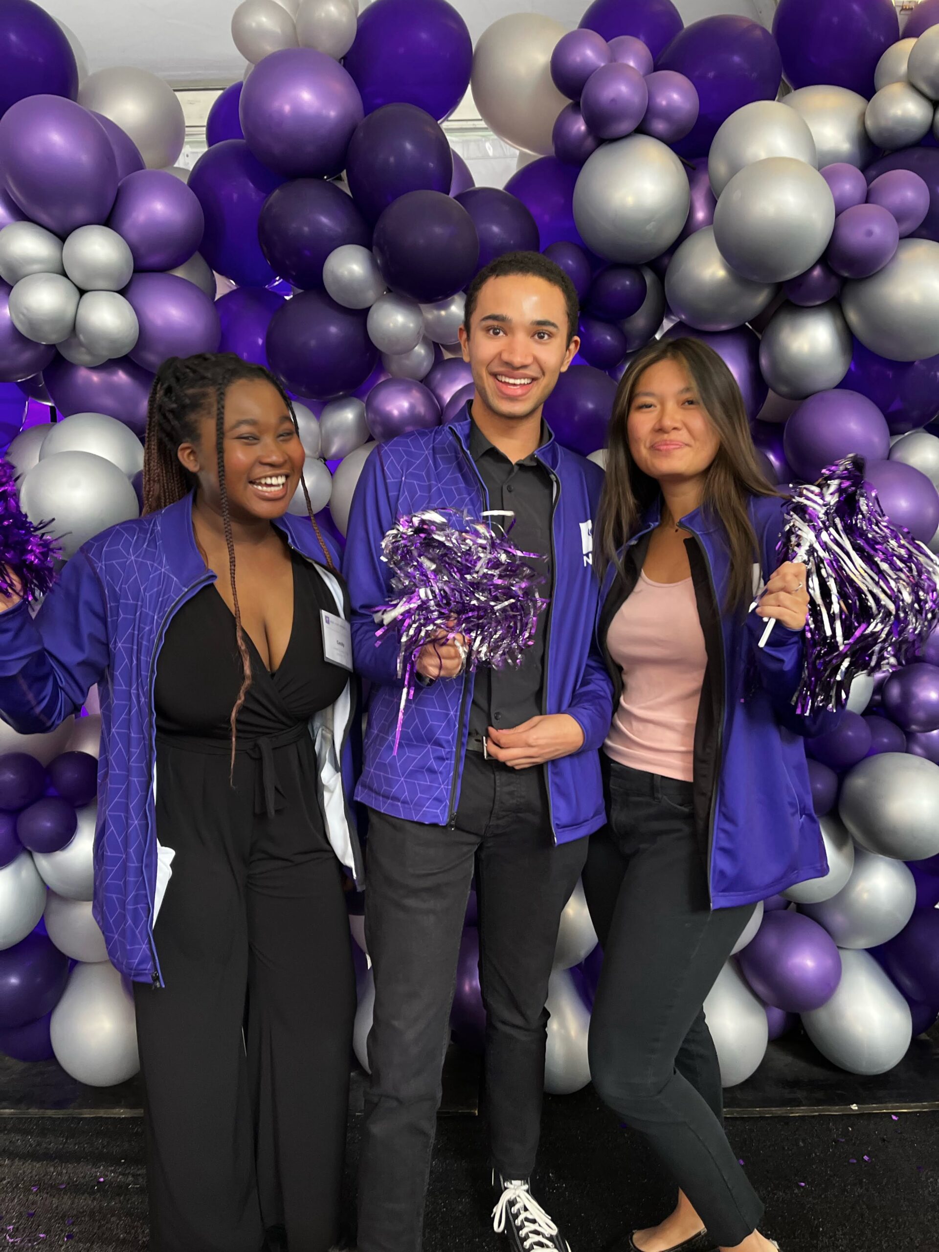 Three friends wearing purple jackets and surrounded by purple and silver balloons smile at a work event.