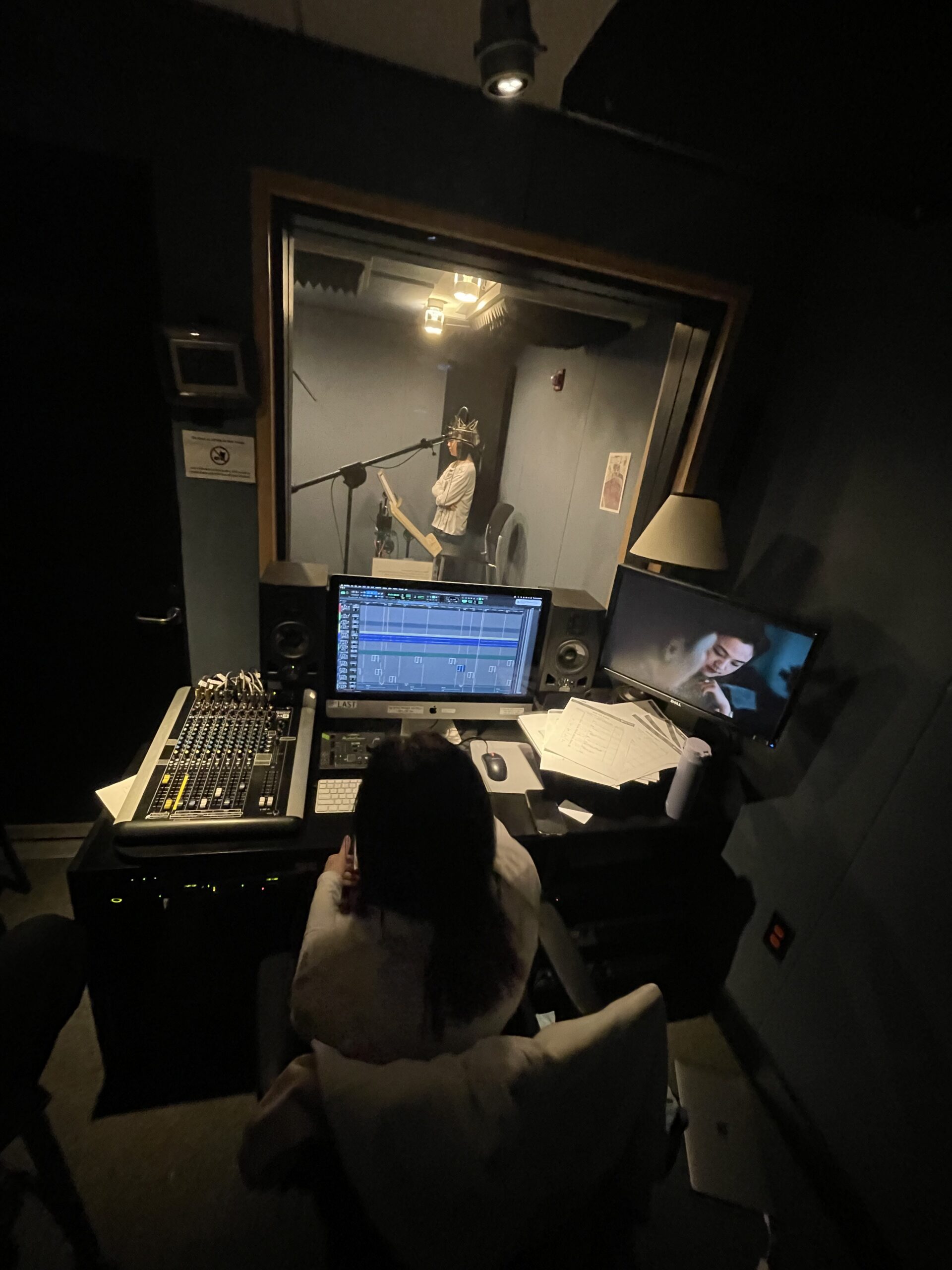 Behind the scenes of a sound mix craft course in an automated dialogue replacement room. One student records in a booth while another produces the sound.