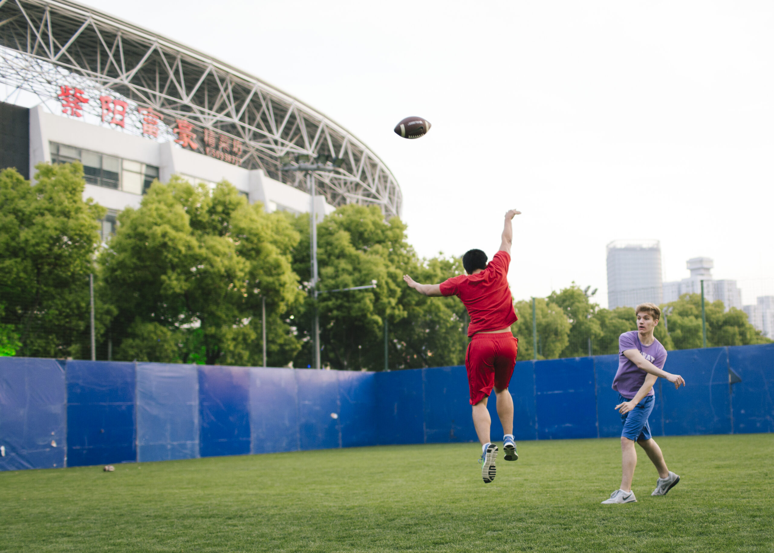 Two students playing football on a grassy field. There’s a large stadium and city buildings in the background.