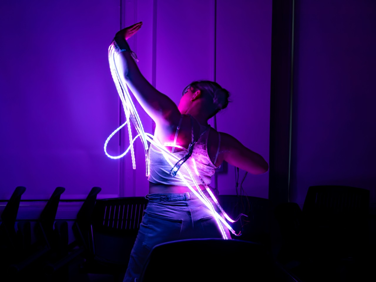 A person performing a dance or movement with illuminated LED light strips wrapped around their body in a dark room, creating a vibrant display of purple and blue light.
