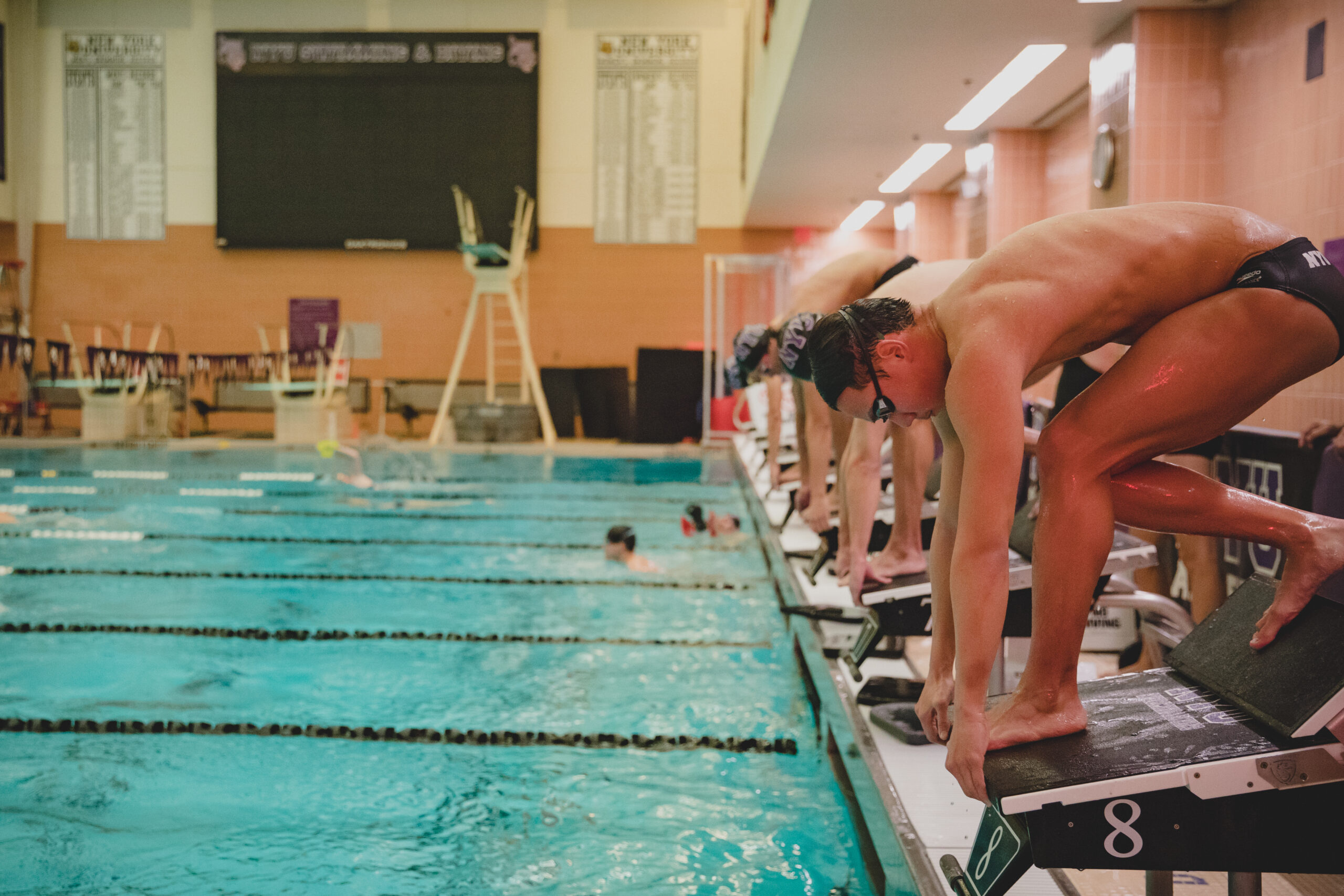 Male-presenting swimmers are poised on starting blocks, ready to dive into the pool for a race.