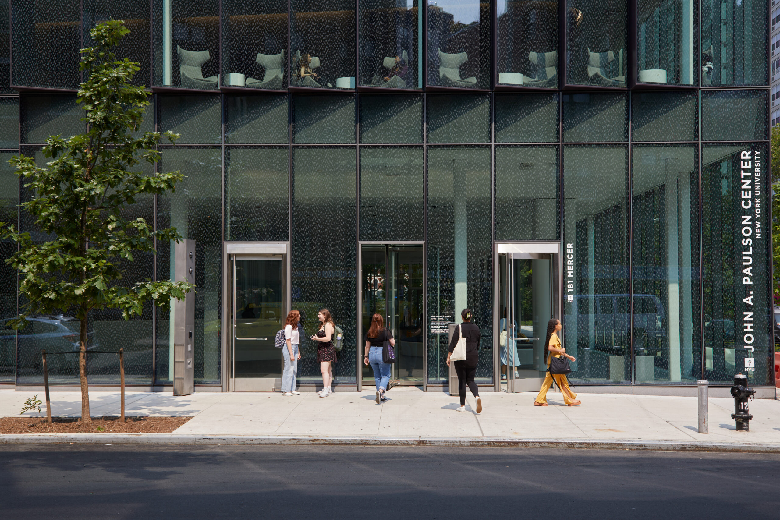 People entering and exiting the John A. Paulson Center on NYU’s campus in New York City. The building has a modern glass facade, and there are a few people standing and chatting outside the entrance.