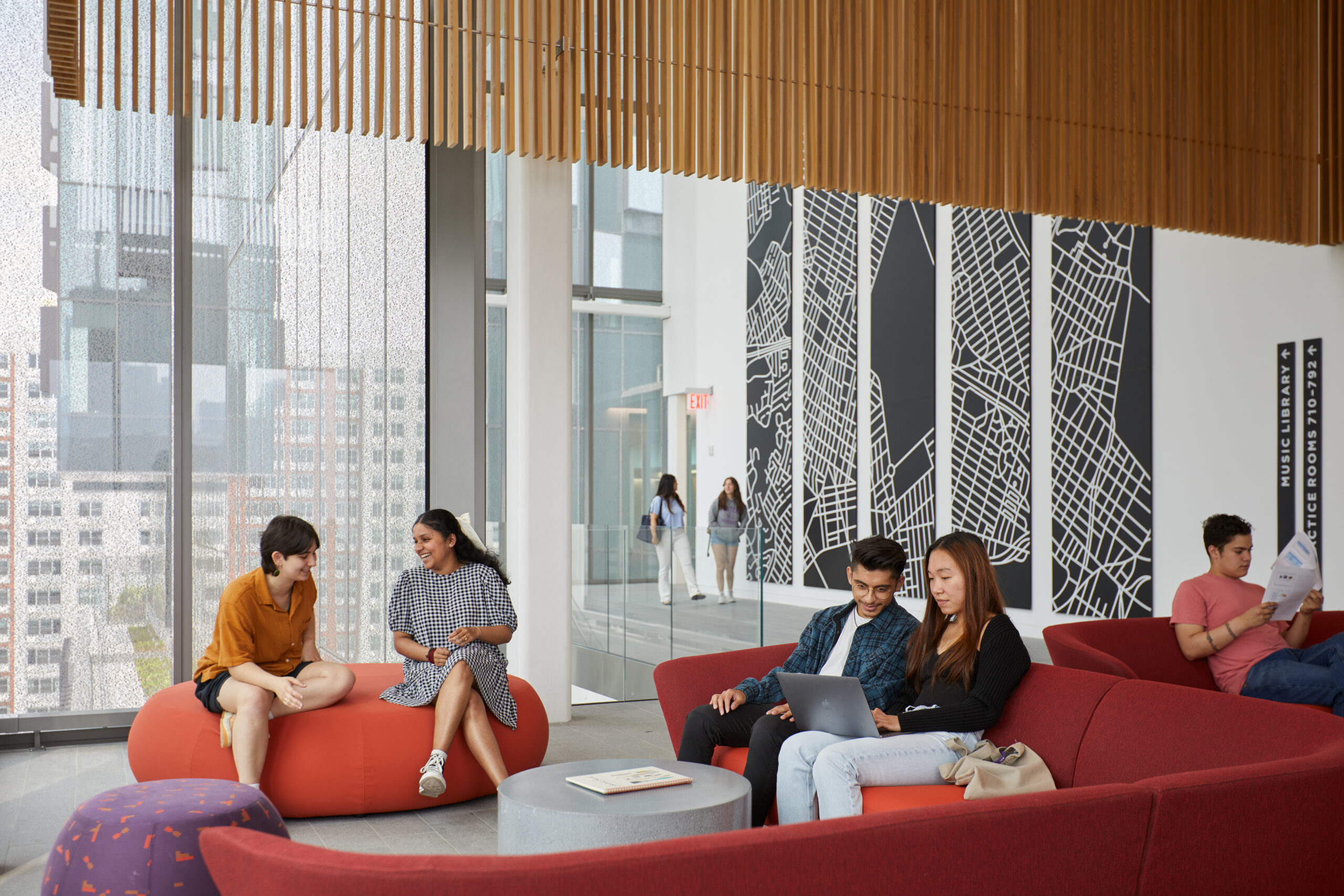 Students sit on vibrant, modern furniture in a well-lit common area, engaging in conversations, working on a laptop, and reading.