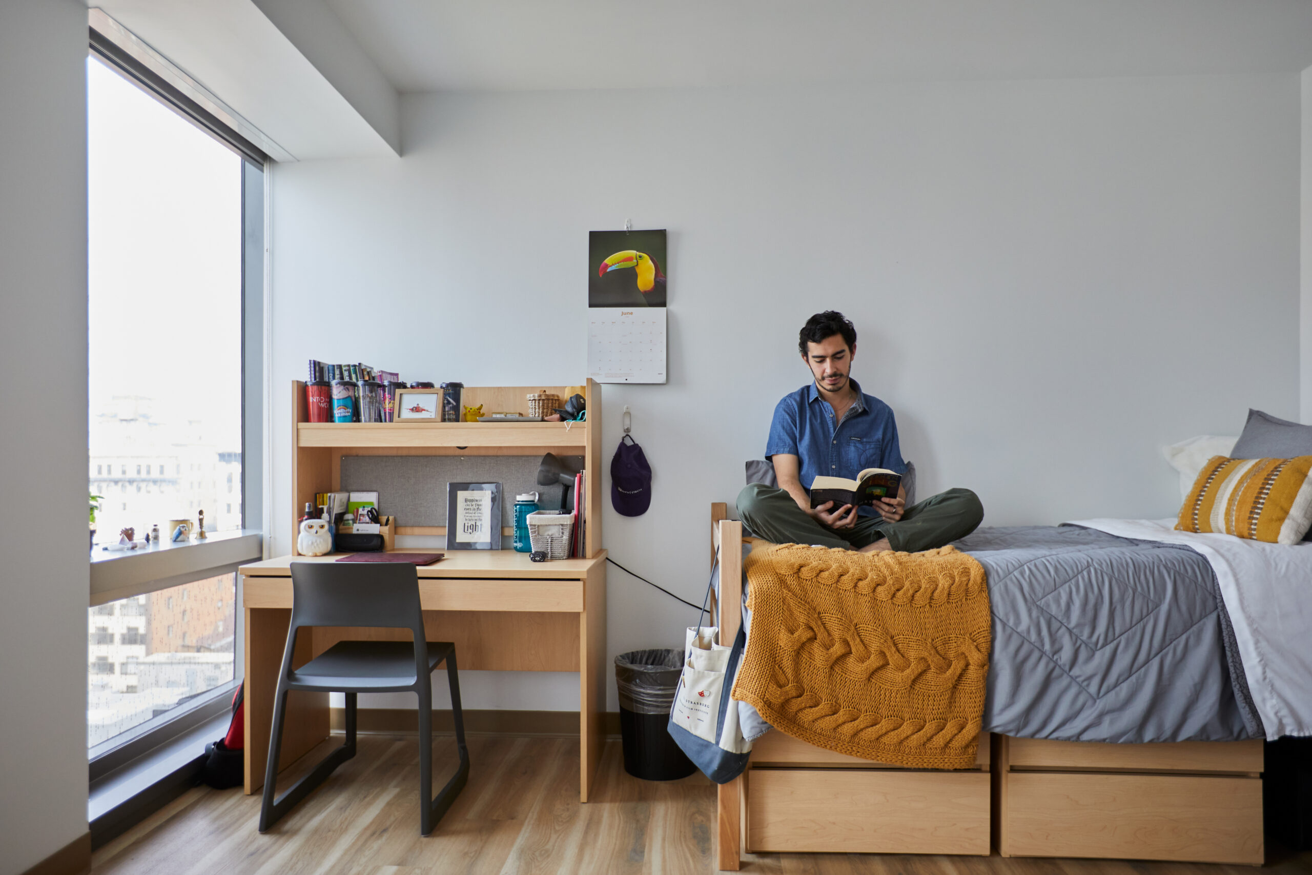 A male-presenting student sits cross-legged on their bed, reading a book in a bright and tidy dorm room.