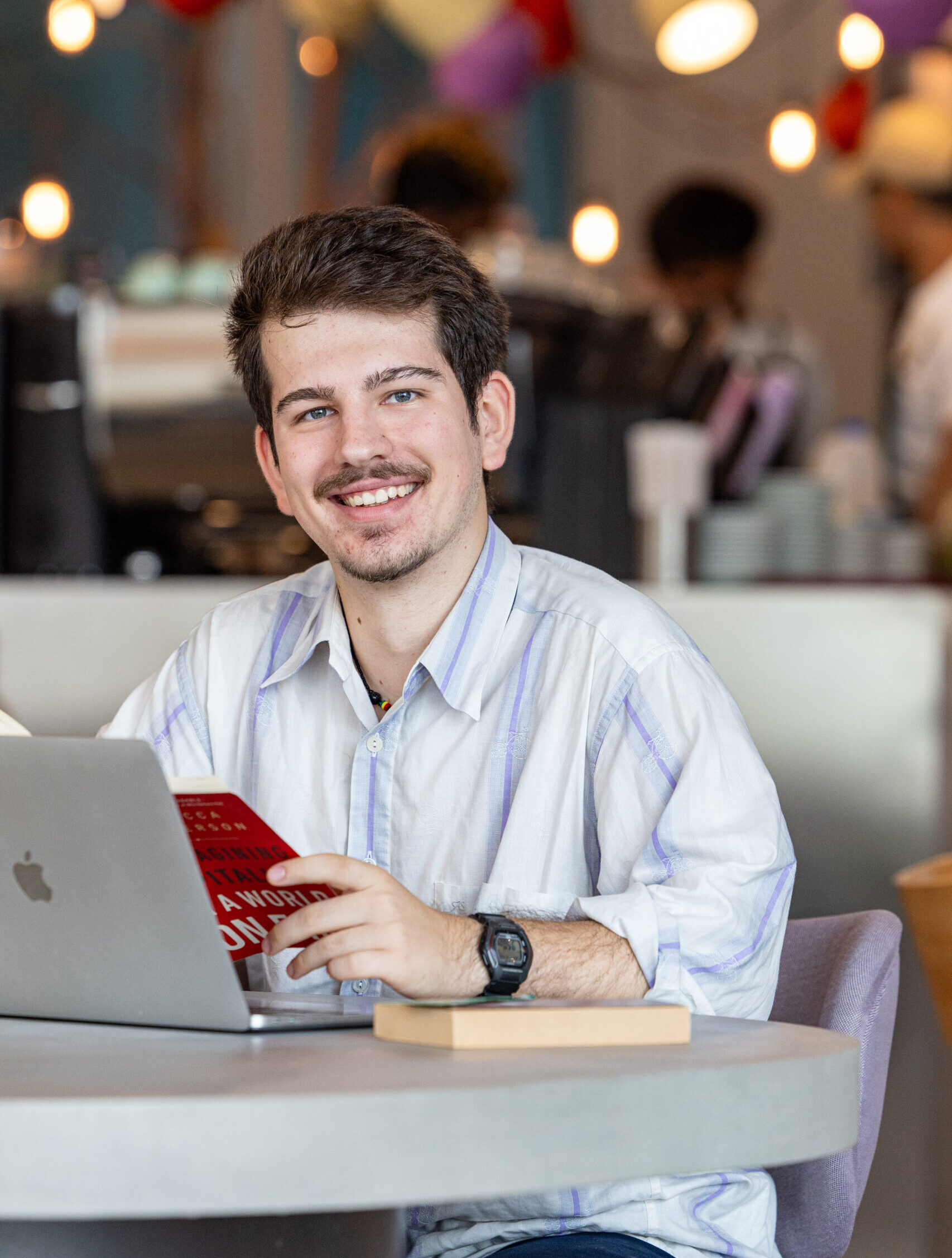 Stefan Mitikj, an NYU Abu Dhabi alum, sits in front of a laptop with a book in his hands.