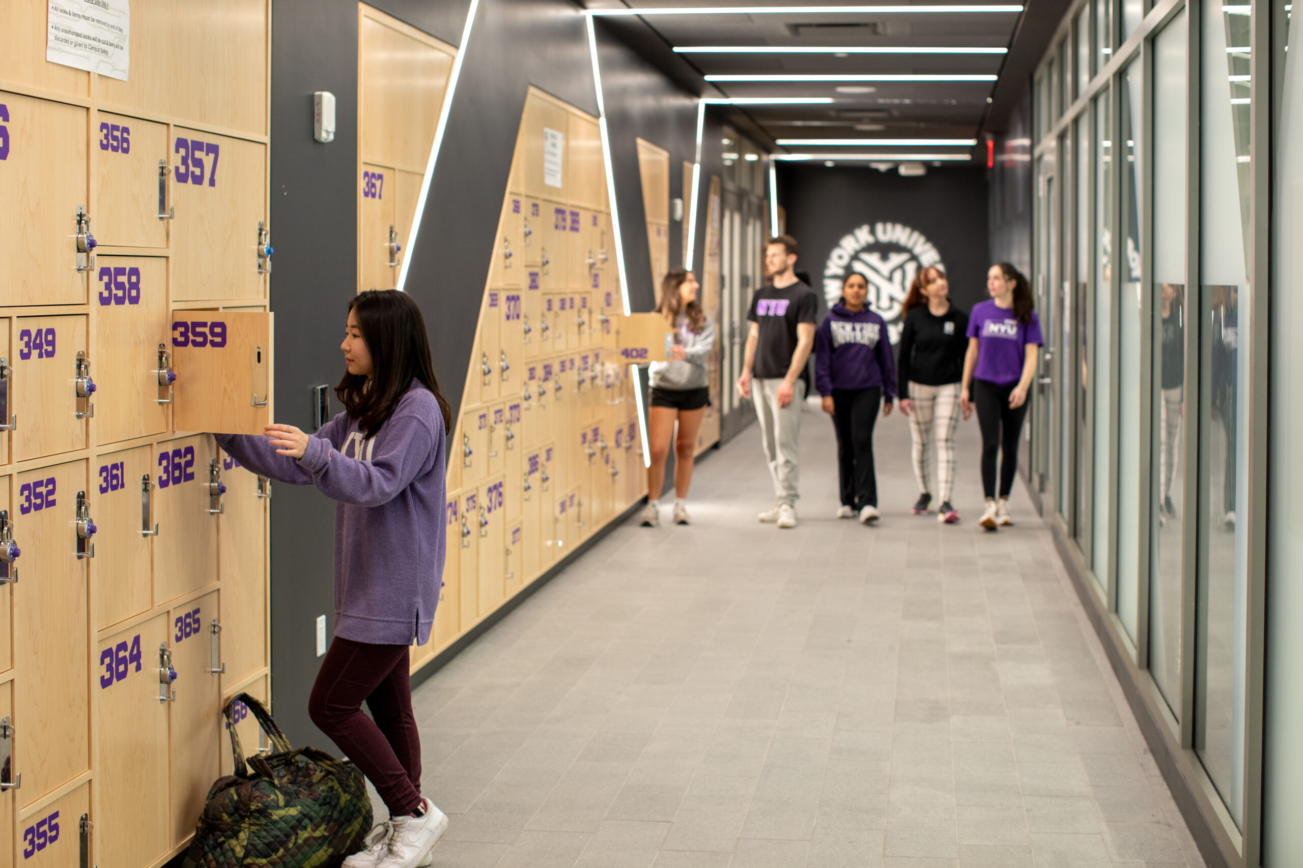 A student wearing a purple sweater places items into a locker numbered 359 in a hallway lined with wooden lockers. In the background, a group of students wearing New York University apparel walks down the hallway.