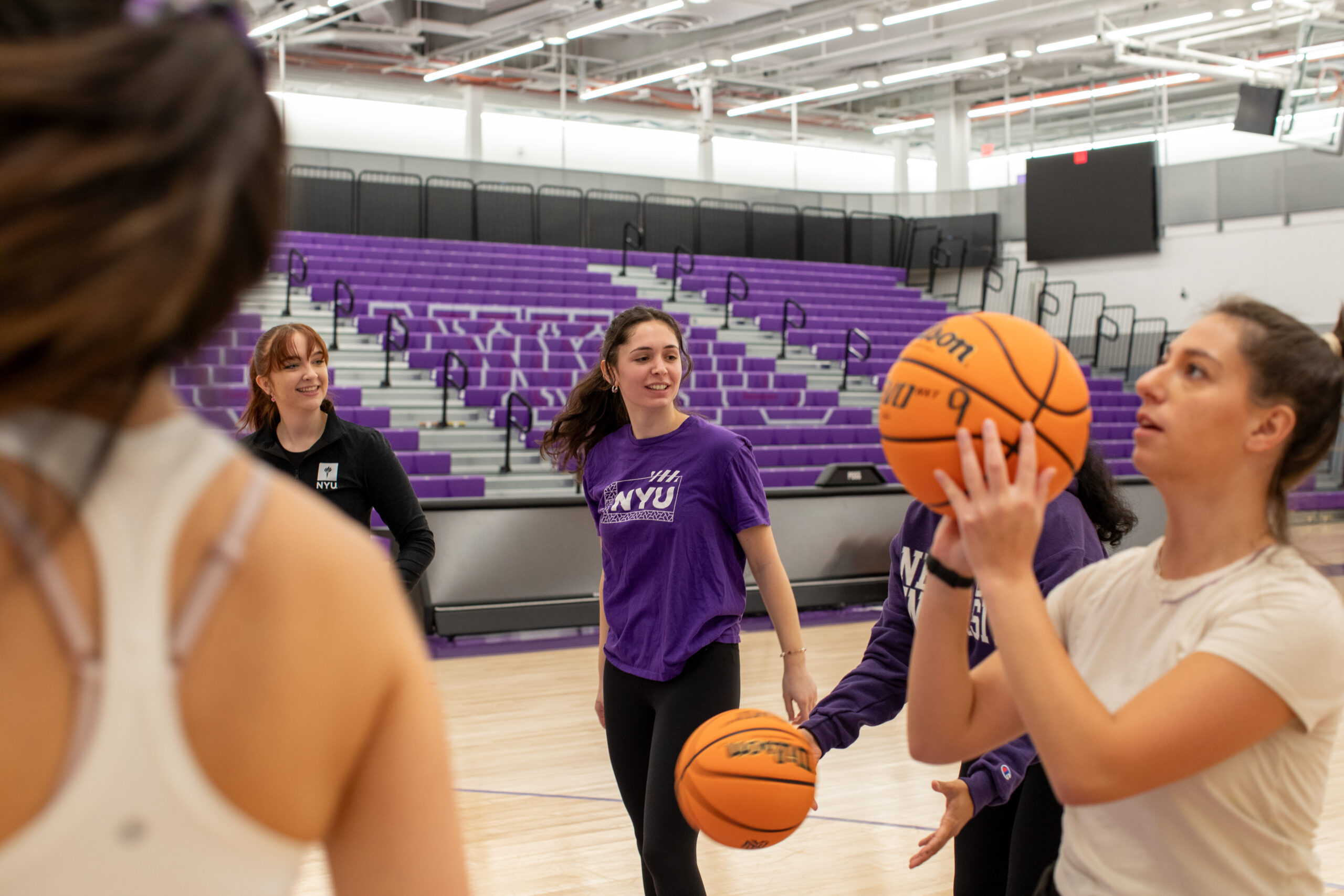 A group of female-presenting students practicing basketball in an indoor gym. One student prepares to shoot while others look on.