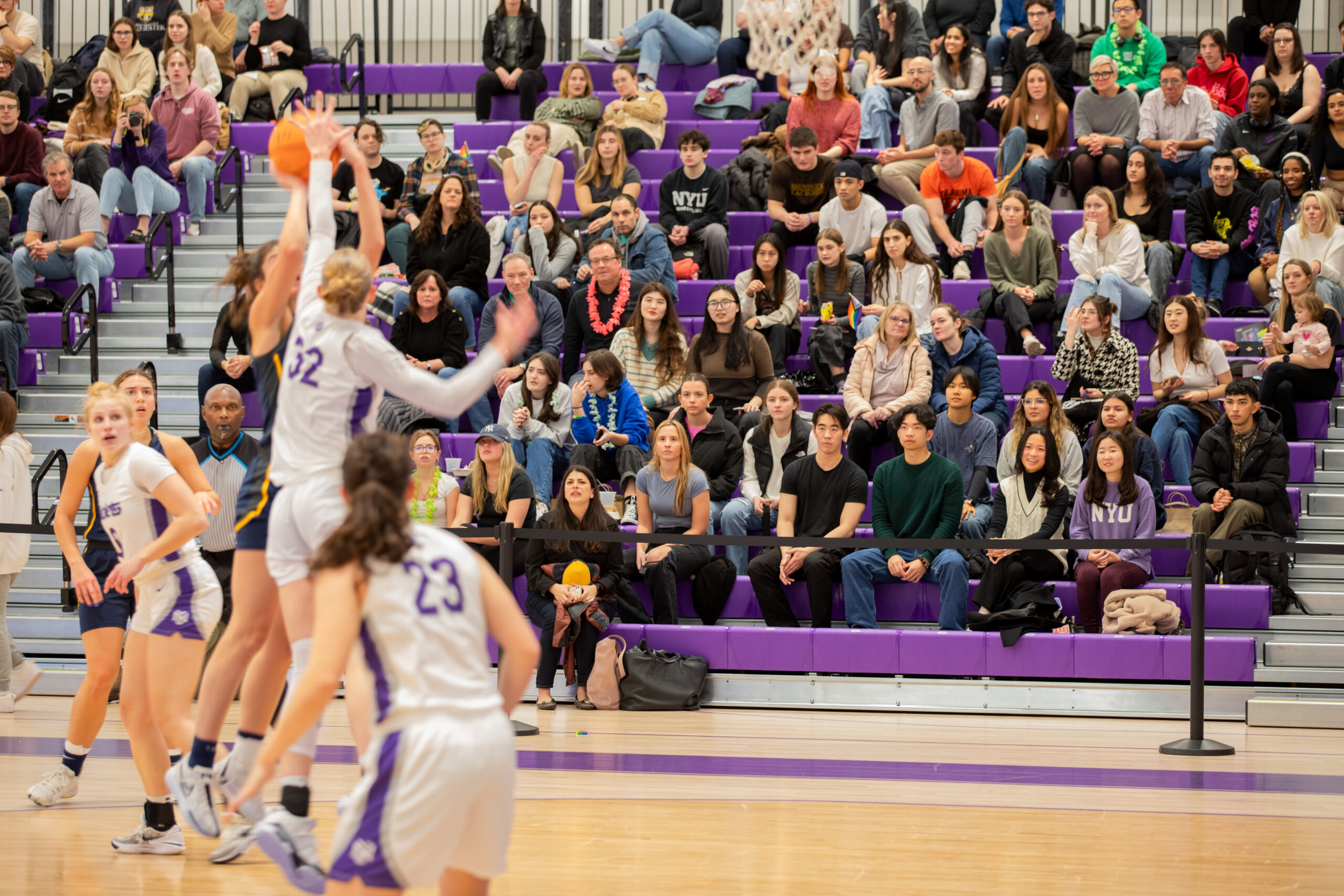 A women’s basketball game is in action with a player shooting the ball to try to make a basket. The defense player attempts to block the shot. The stands in the background are filled with spectators.