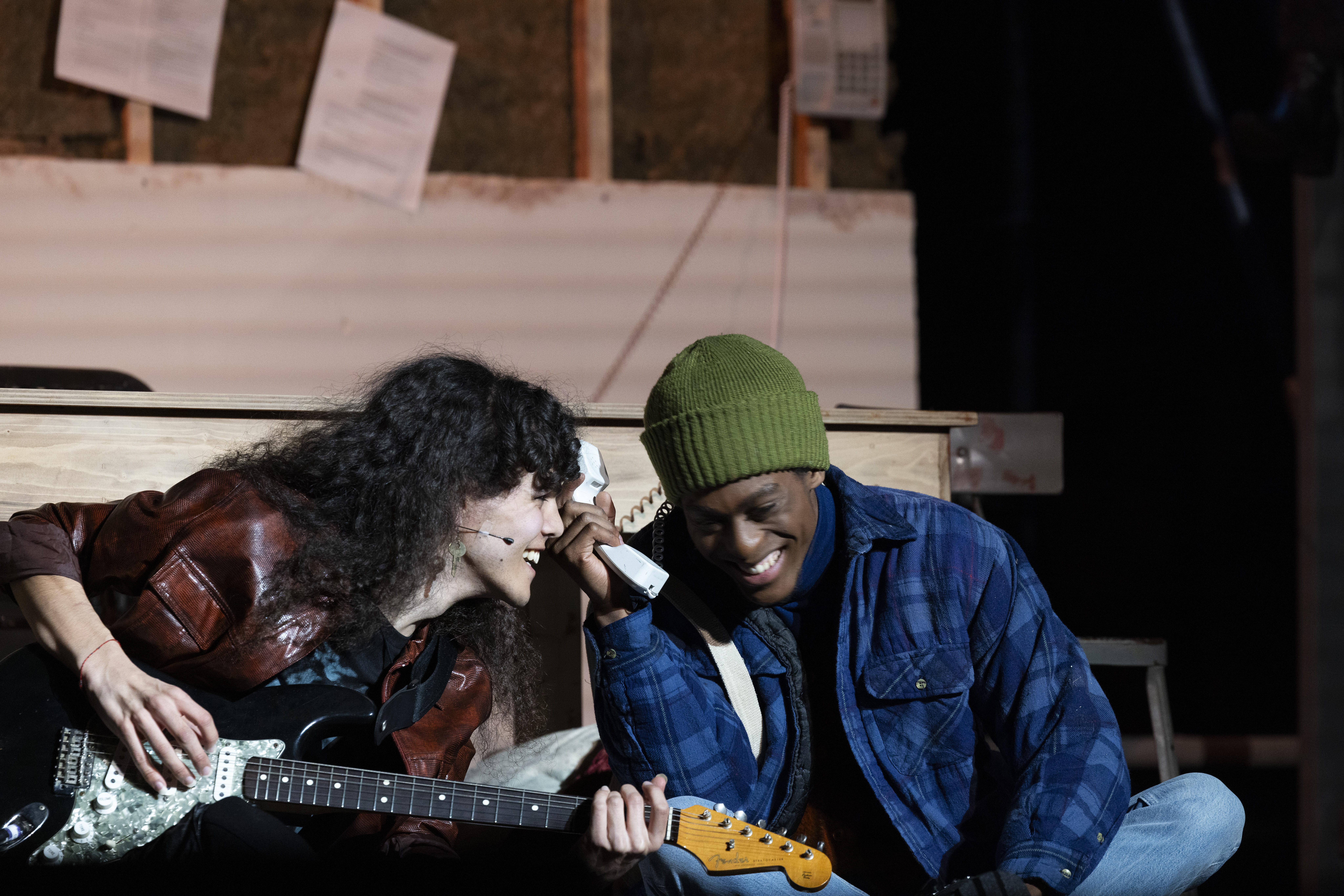 Two students sit close together, one plays an electric guitar while the other holds a telephone receiver, during a production of “Rent.”