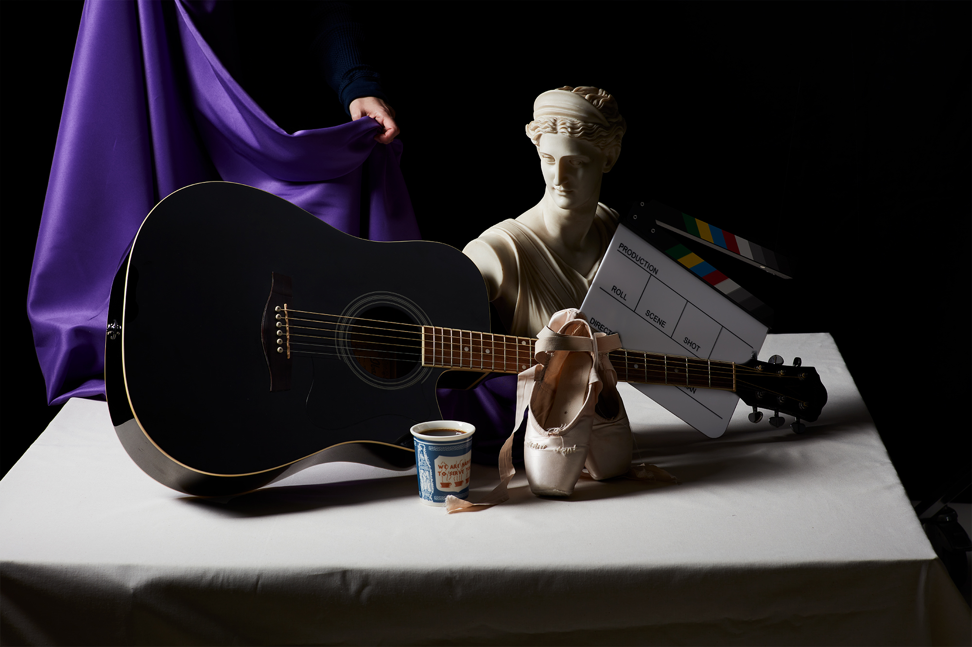 A still life arrangement featuring a guitar, coffee cup, ballet shoes, clapboard, and statue bust, with a hand adjusting the violet tablecloth from the left side.