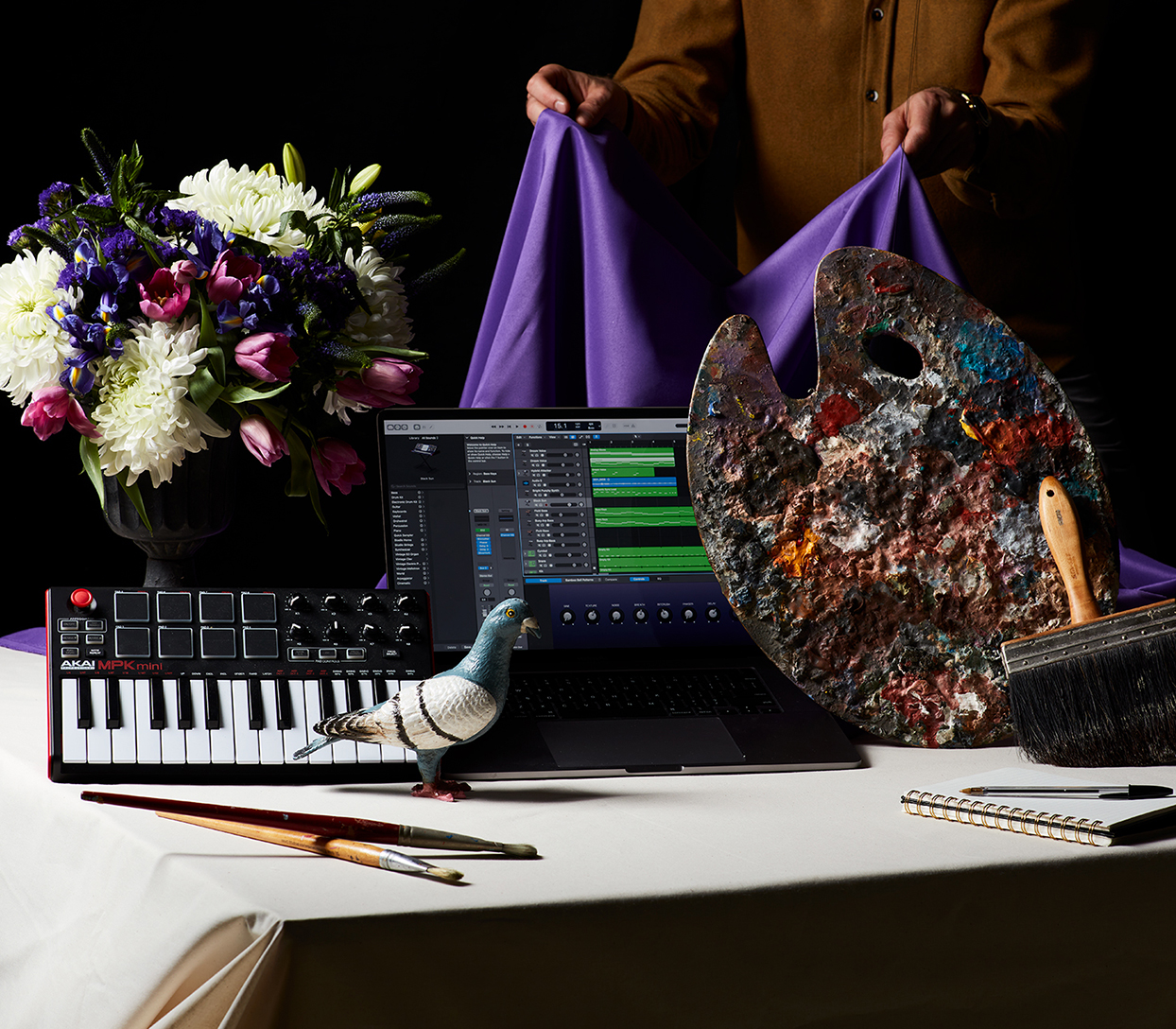 A creative still life composition featuring a MIDI keyboard, a pigeon statue, a painter’s palette, paintbrushes, flowers in a vase, and a laptop displaying music production software. A person holds purple fabric in the background.