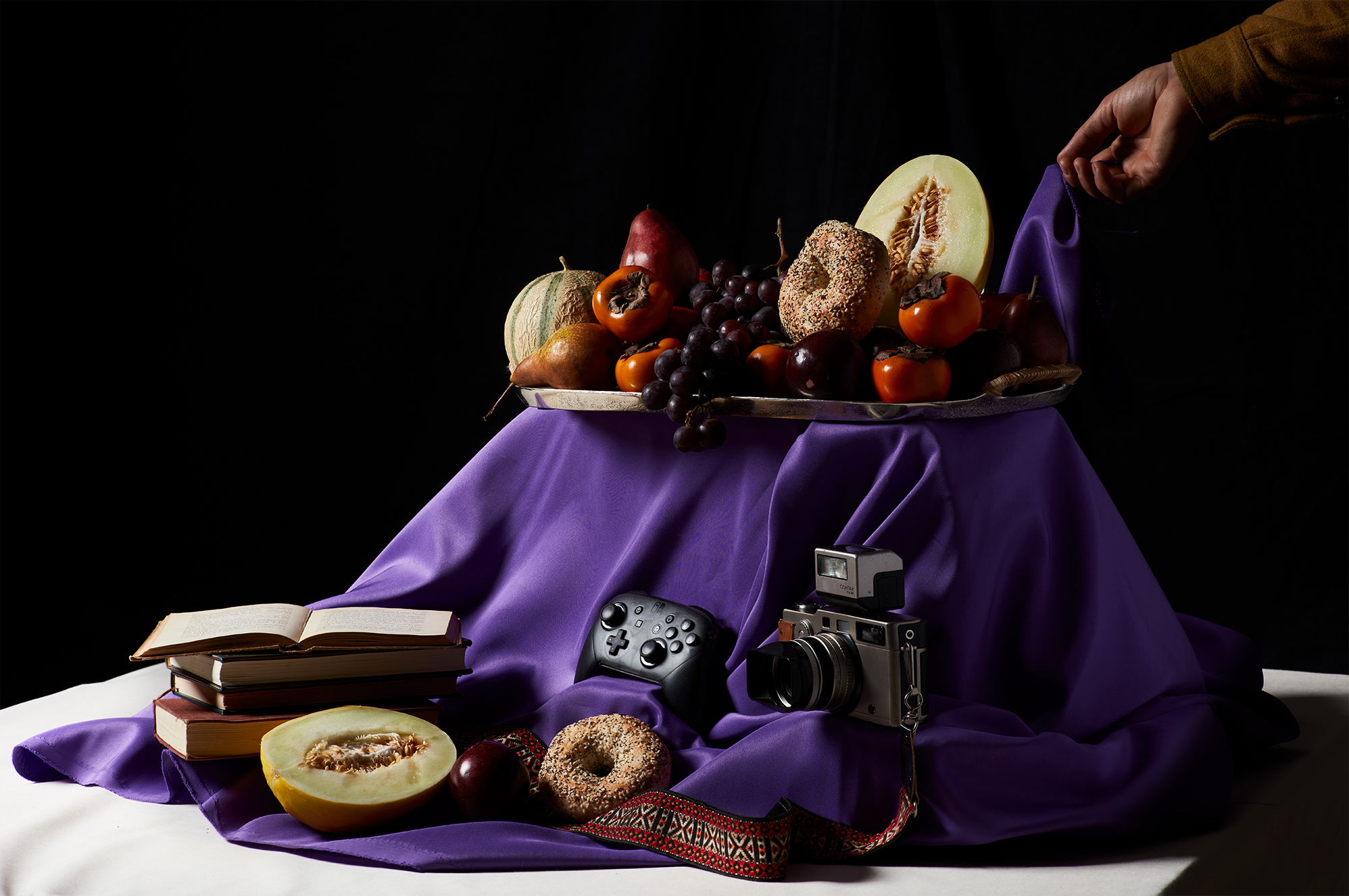 A still life arrangement featuring a variety of fruits, bagels, and books on a purple cloth, alongside a vintage camera and a video game controller. A person’s hand adjusts the cloth from the right side.