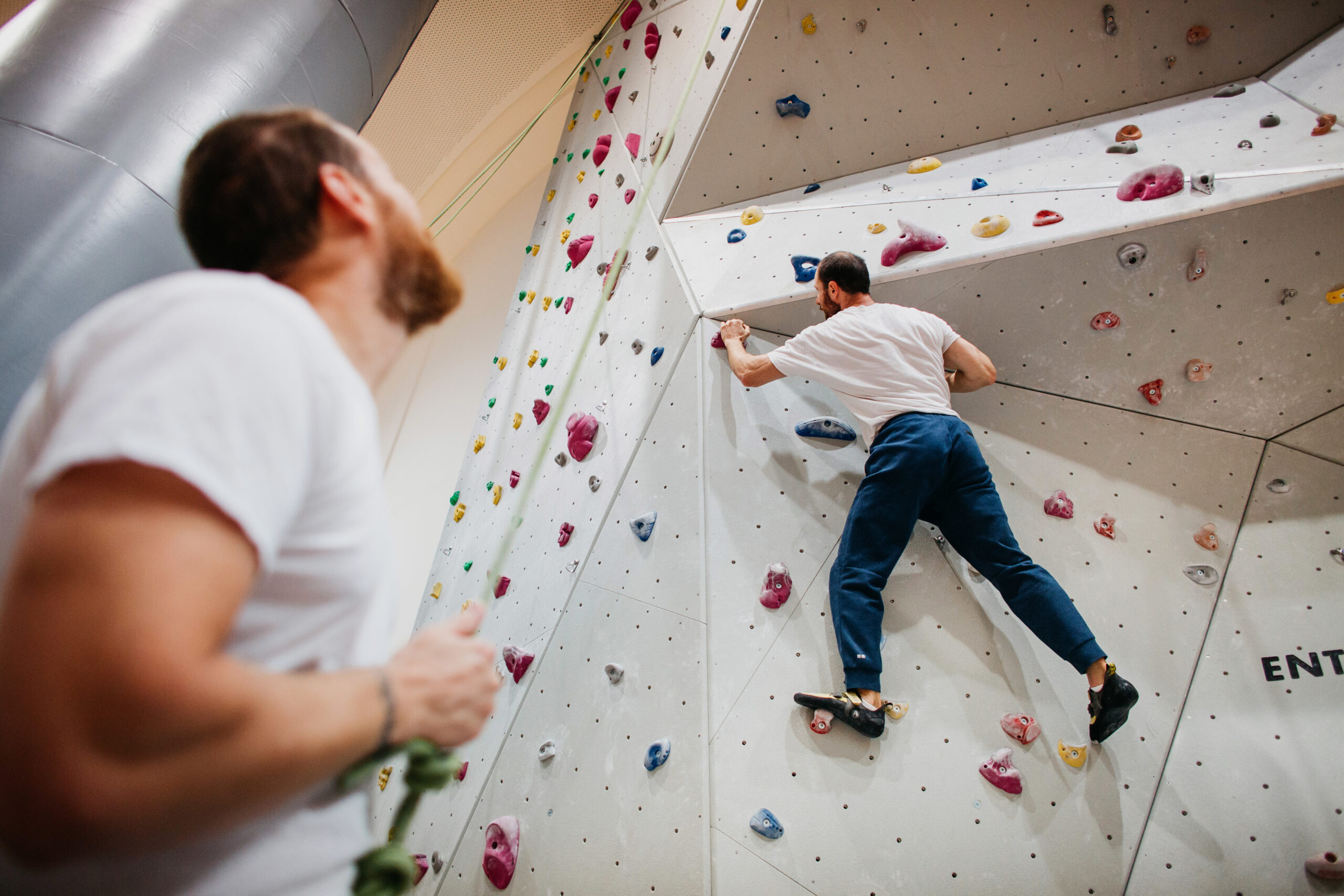 One male-presenting student free-climbs an indoor rock climbing wall while another male-presenting student belays.