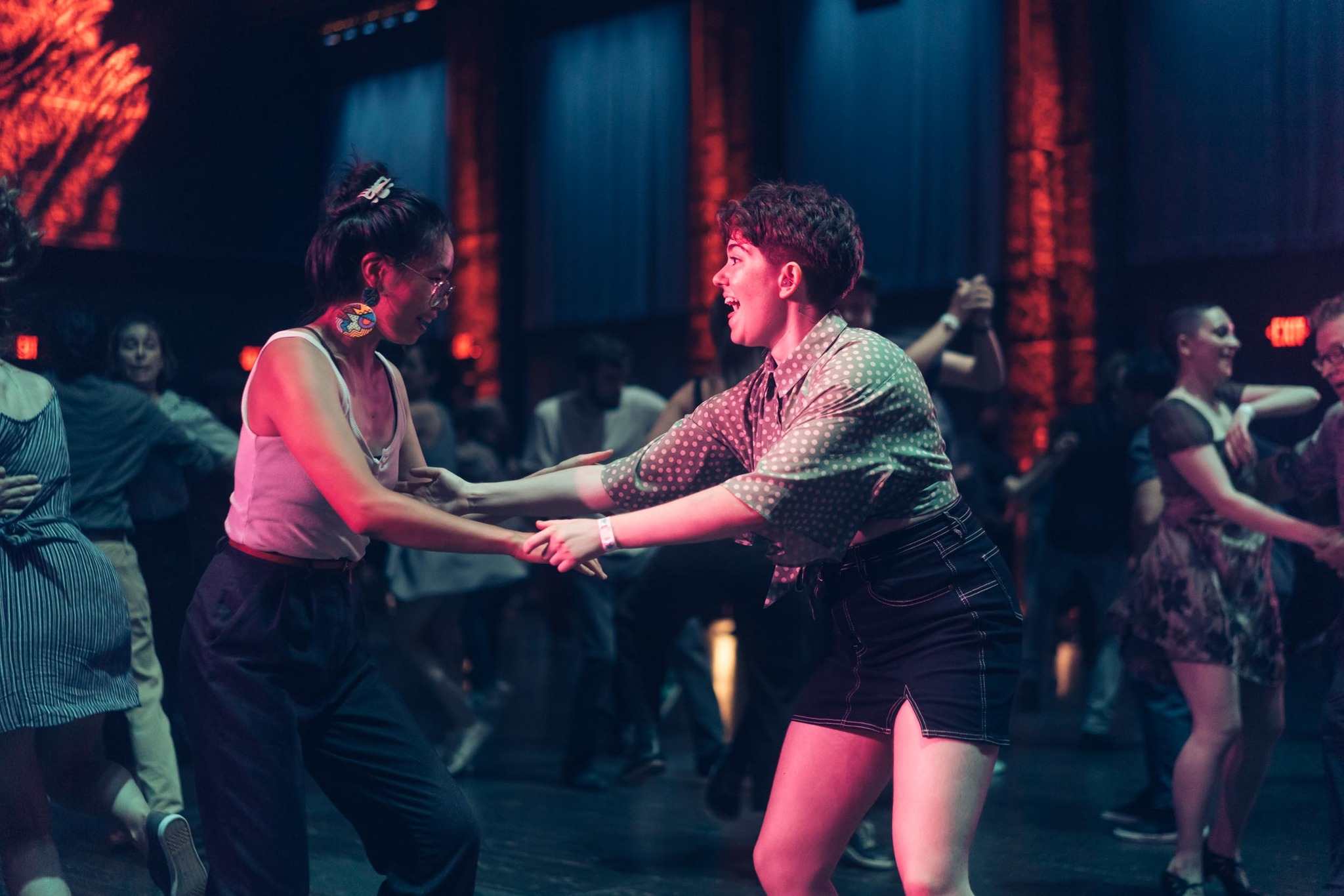 Two people dance together in a dimly lit, colorful setting surrounded by other dancing pairs.