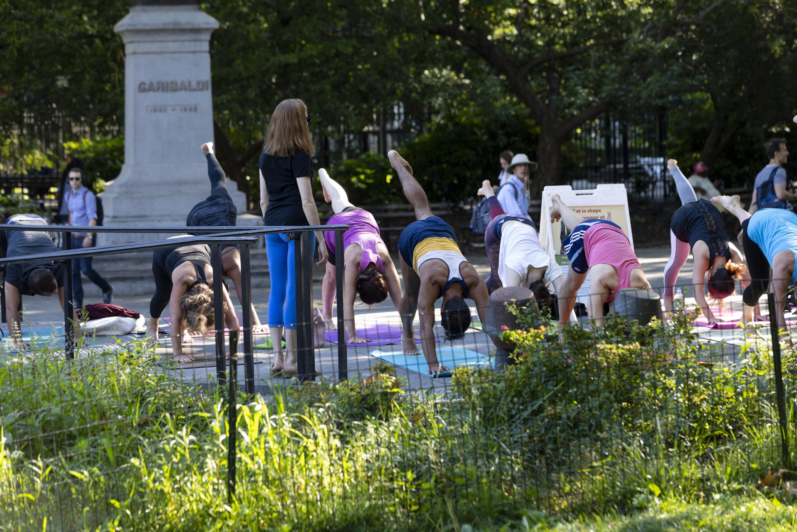 A group of people practicing yoga outdoors in a park. Each person is in three-legged downward dog pose.