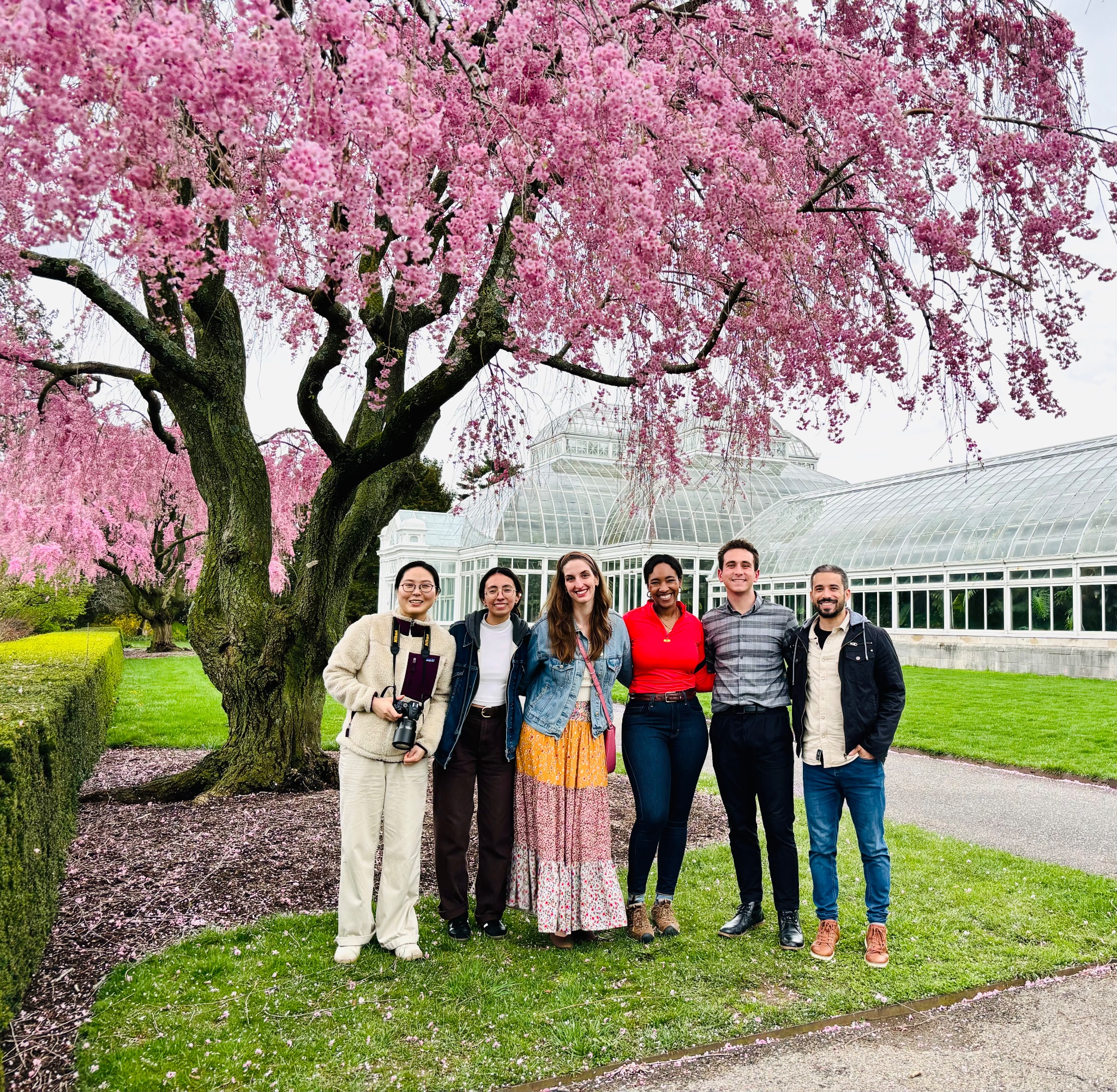 Professor Joyce Onyenedum stands in the middle of a group of researchers she works with in the lab in front of a pink cherry blossom tree outside of a greenhouse.