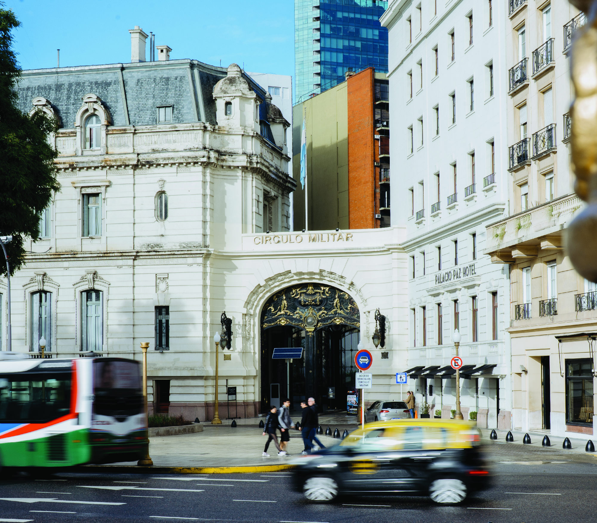 A busy street in front of the historic Círculo Militar building in Buenos Aires, Argentina. A car and bus are in motion and pedestrians cross the street.