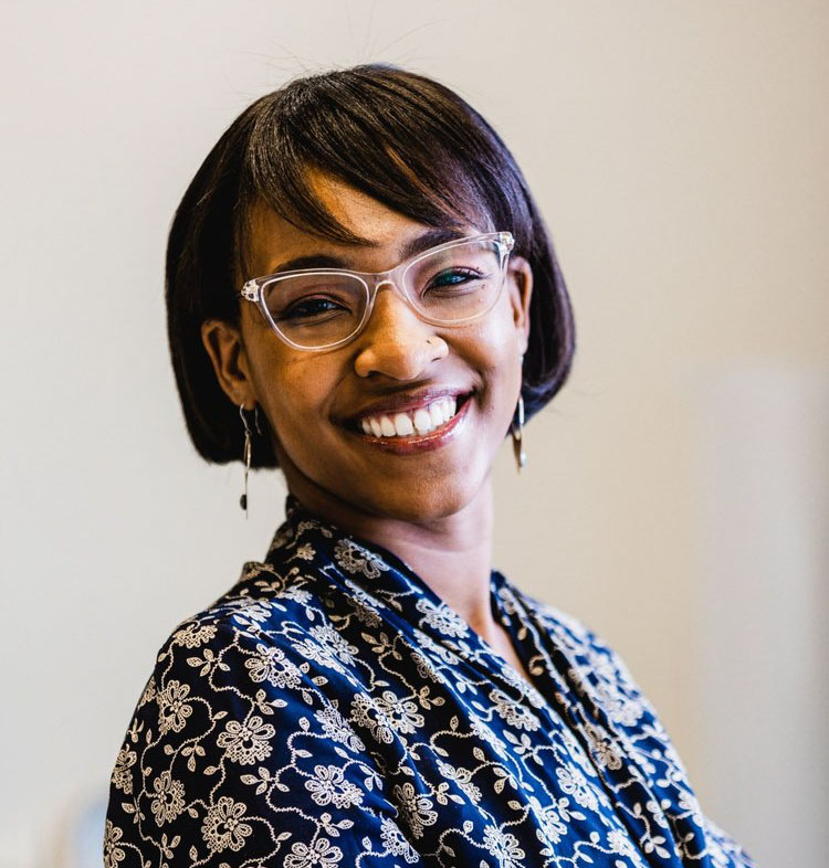 NYU professor Joyce Onyenedum studies plant diversity. She is a person of color with short dark hair and bangs. She is wearing clear-framed glasses, dangling earrings, and a navy shirt with a floral pattern.