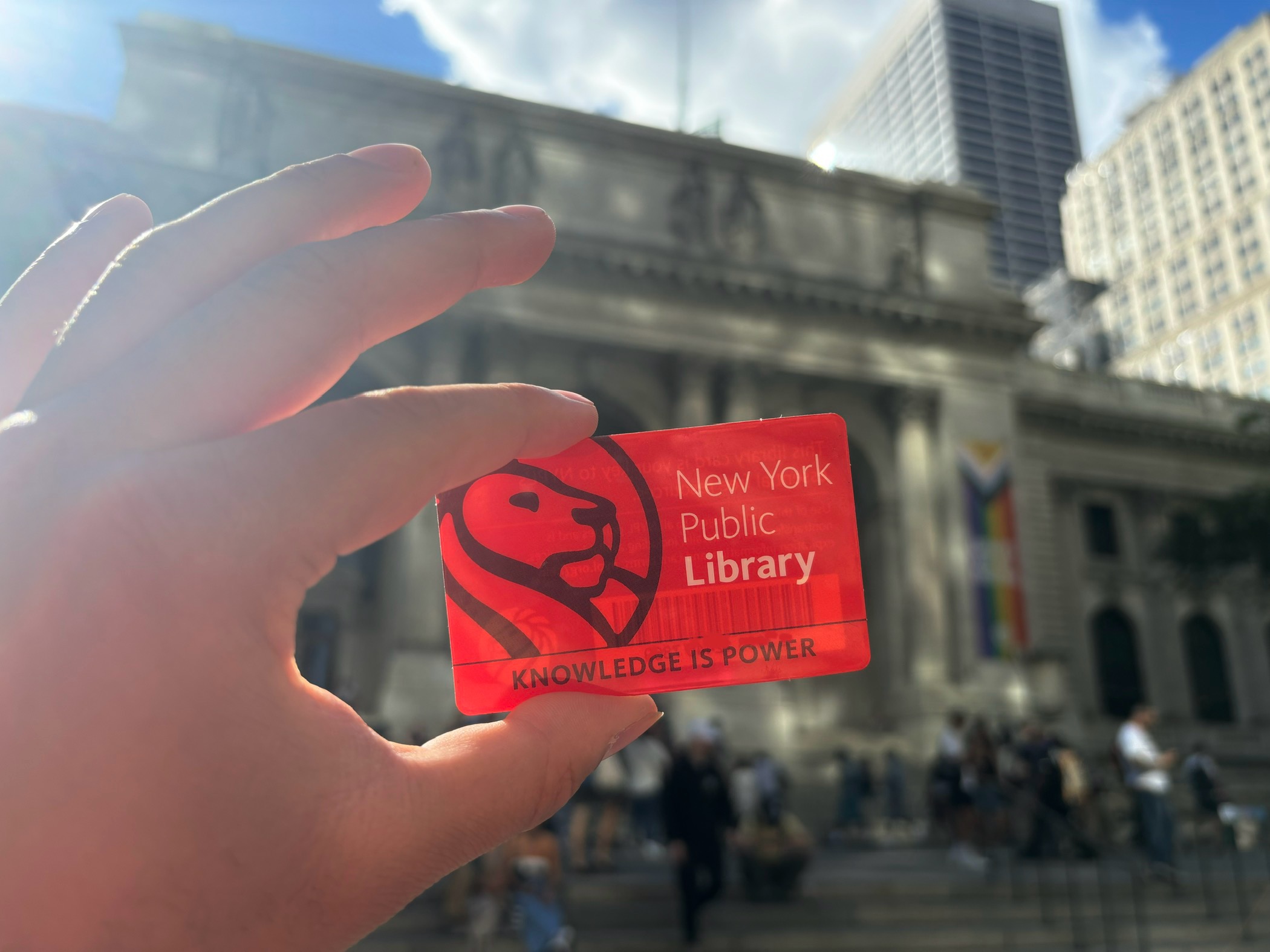A hand holding a New York Public Library card with the main library branch out of focus in the background.