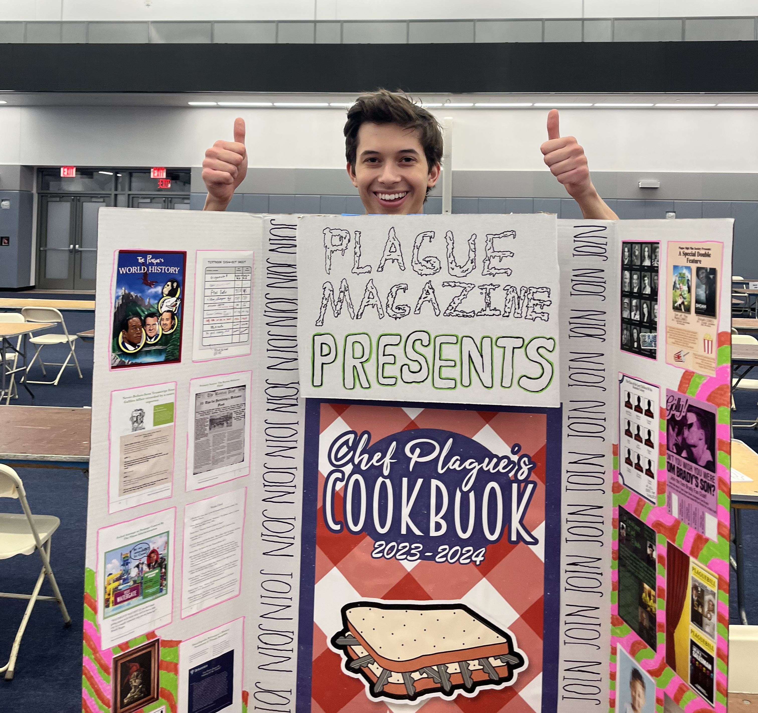 The author, a male-presenting NYU student, stands behind a trifold poster promoting “The Plague,” NYU’s only intentionally funny student publication.