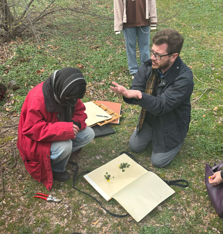 A student in a red jacket and head scarf kneels on the ground, listening to an instructor who is also kneeling and explaining something. Botanical specimens and notebooks lie on the grass between them.