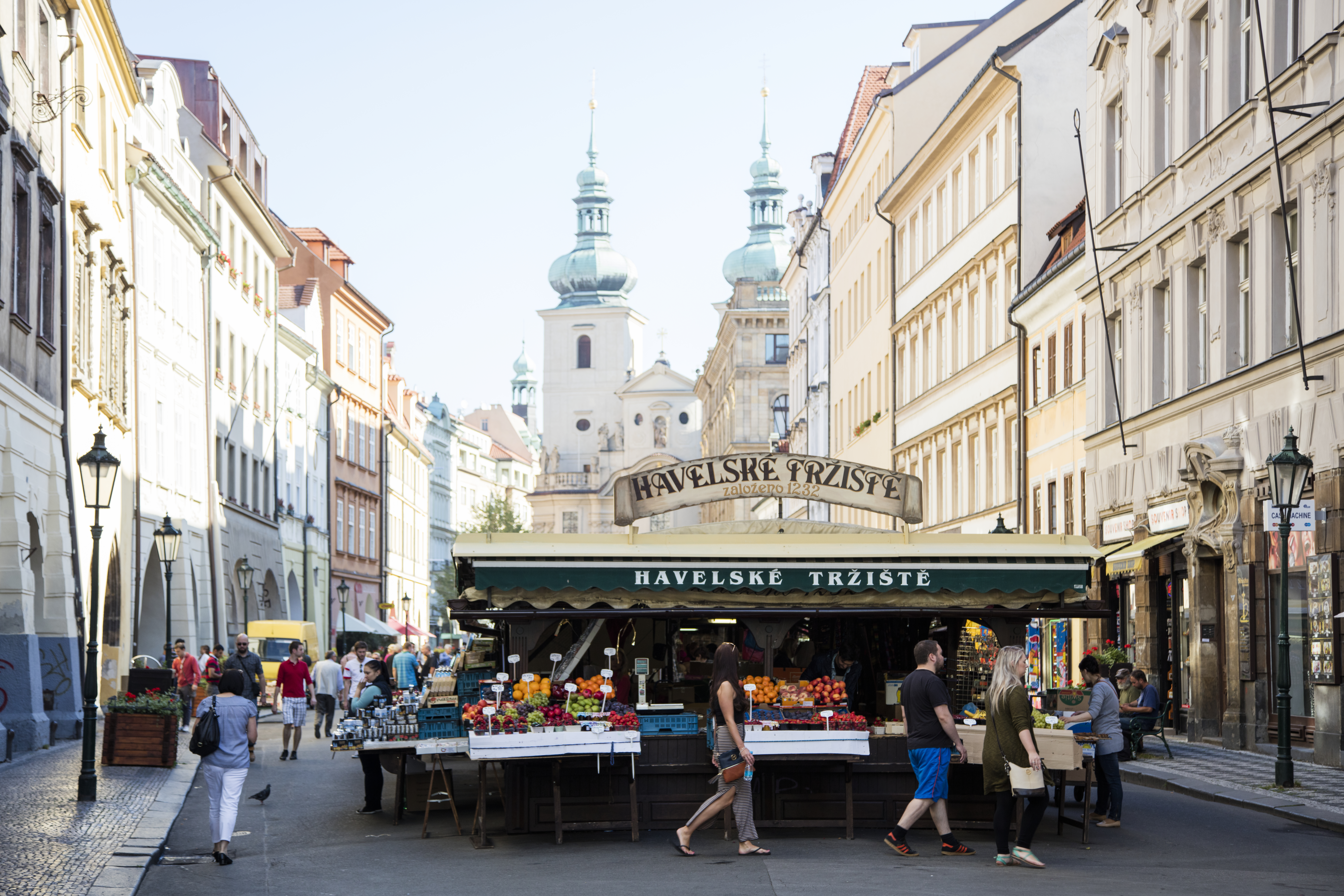 A street market in Prague, flanked by historic buildings and a church in the background. People shop for fruits and vegetables.