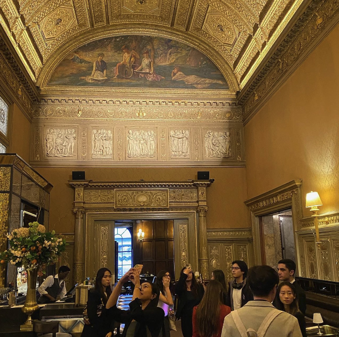 Students and professionals in a luxurious, ornate room with detailed decorations and a large mural on the ceiling, taking photos and admiring the architecture.