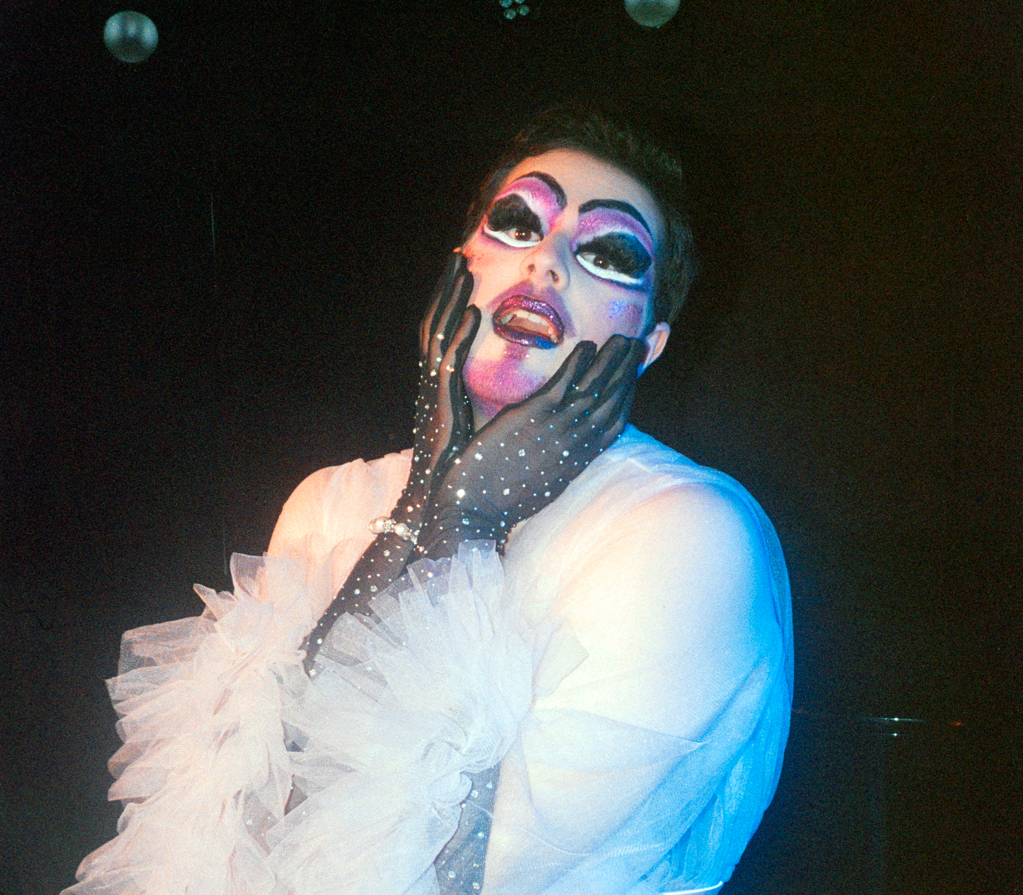 An NYU student, who goes by the name of Toast, dress in drag before a performance. Toast wears bright pink and white makeup, black sparkly gloves, and a white blouse with tulle on the sleeves.