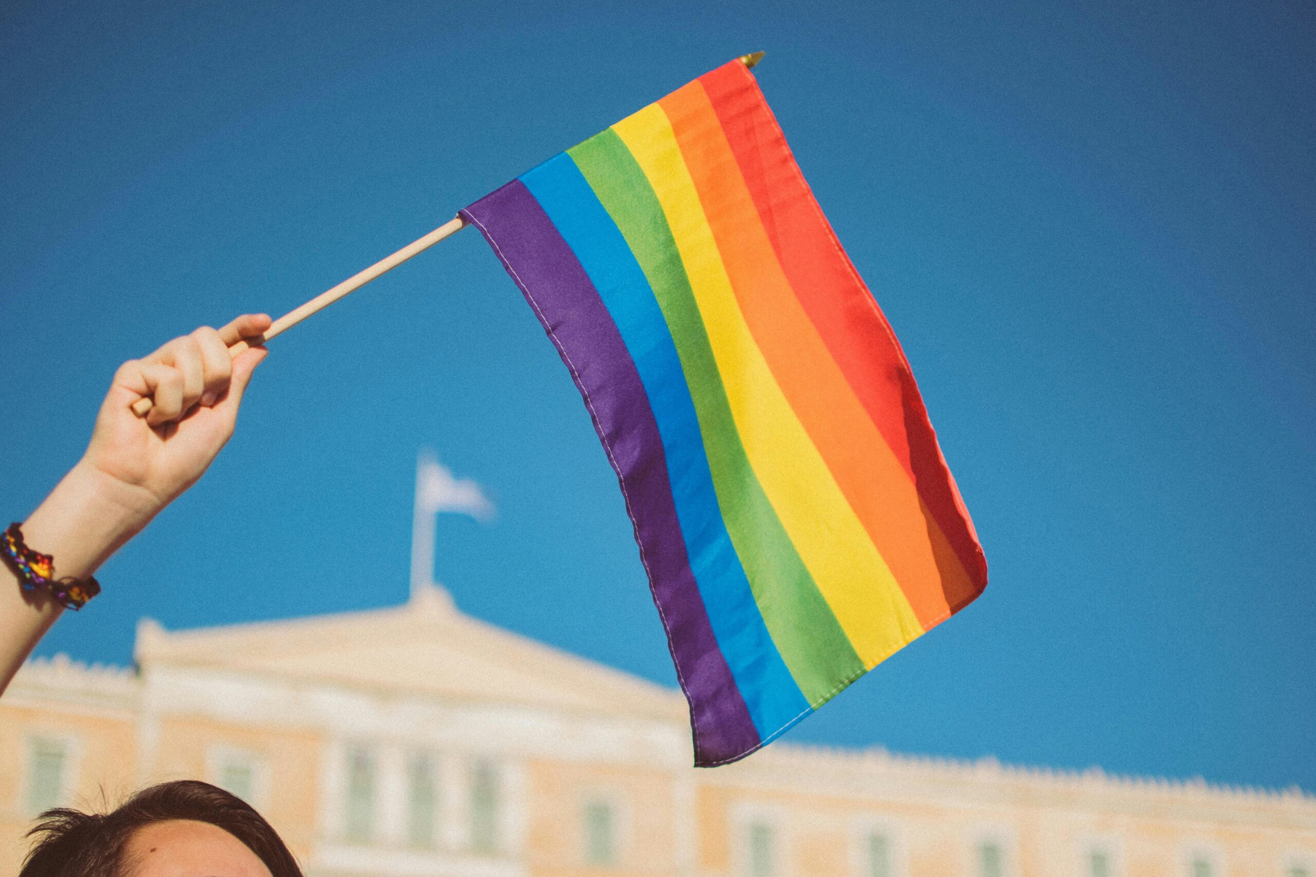 A hand holds a rainbow pride flag against a clear blue sky. A building and flagpole are visible in the background.