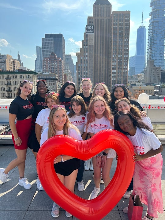 A group of girls smiling on a rooftop with city skyline views holding a heart shaped ballon.