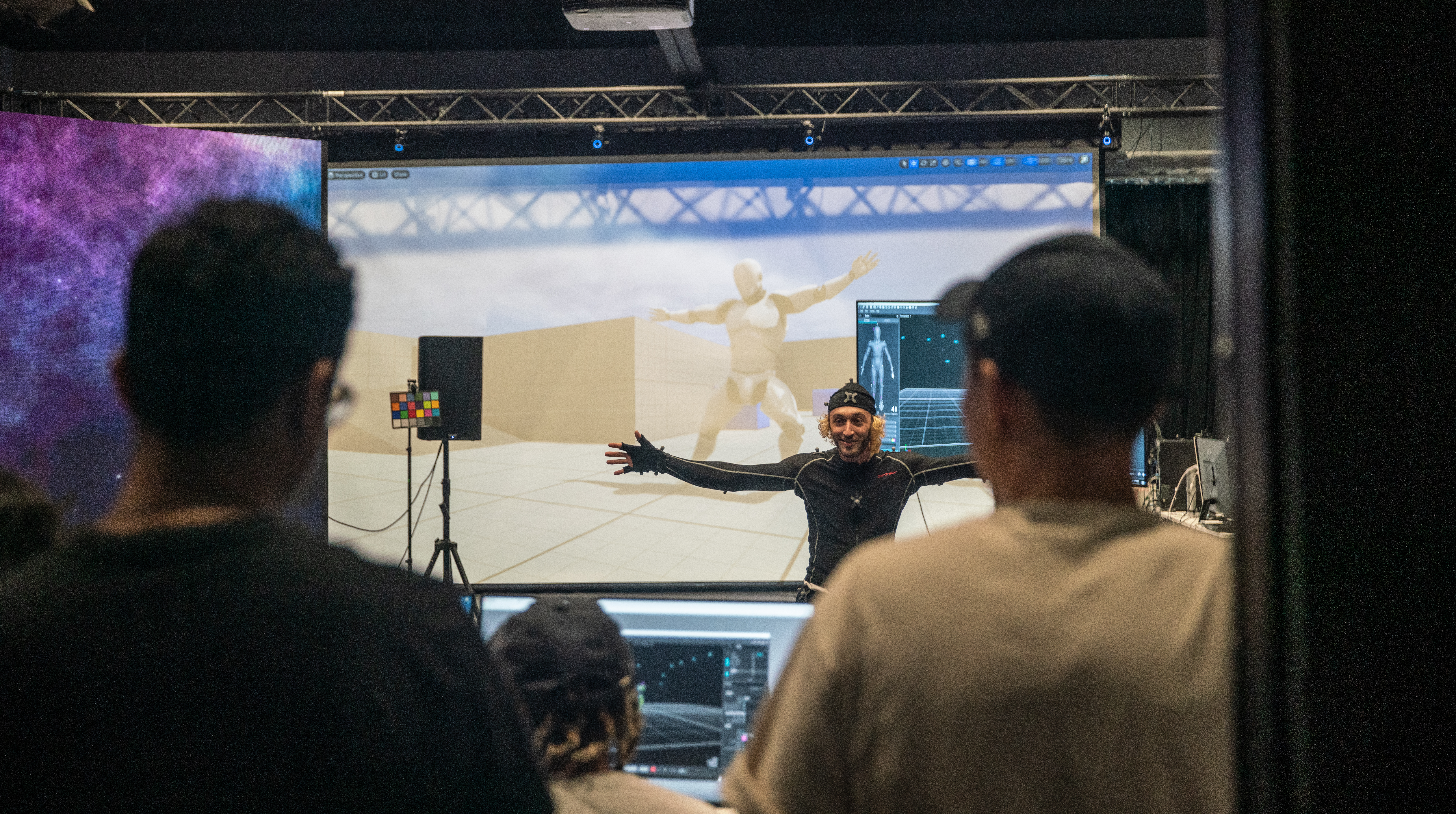 A person wearing motion capture gear moves in front of a group while a 3D character model is displayed on a large screen in the background.