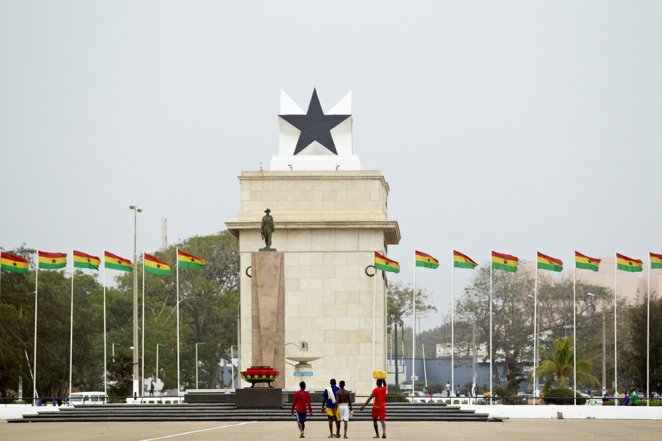 Black Star Gate in Accra, Ghana. Ghanaian flags line the walkway, and a few people walk toward the large monument.