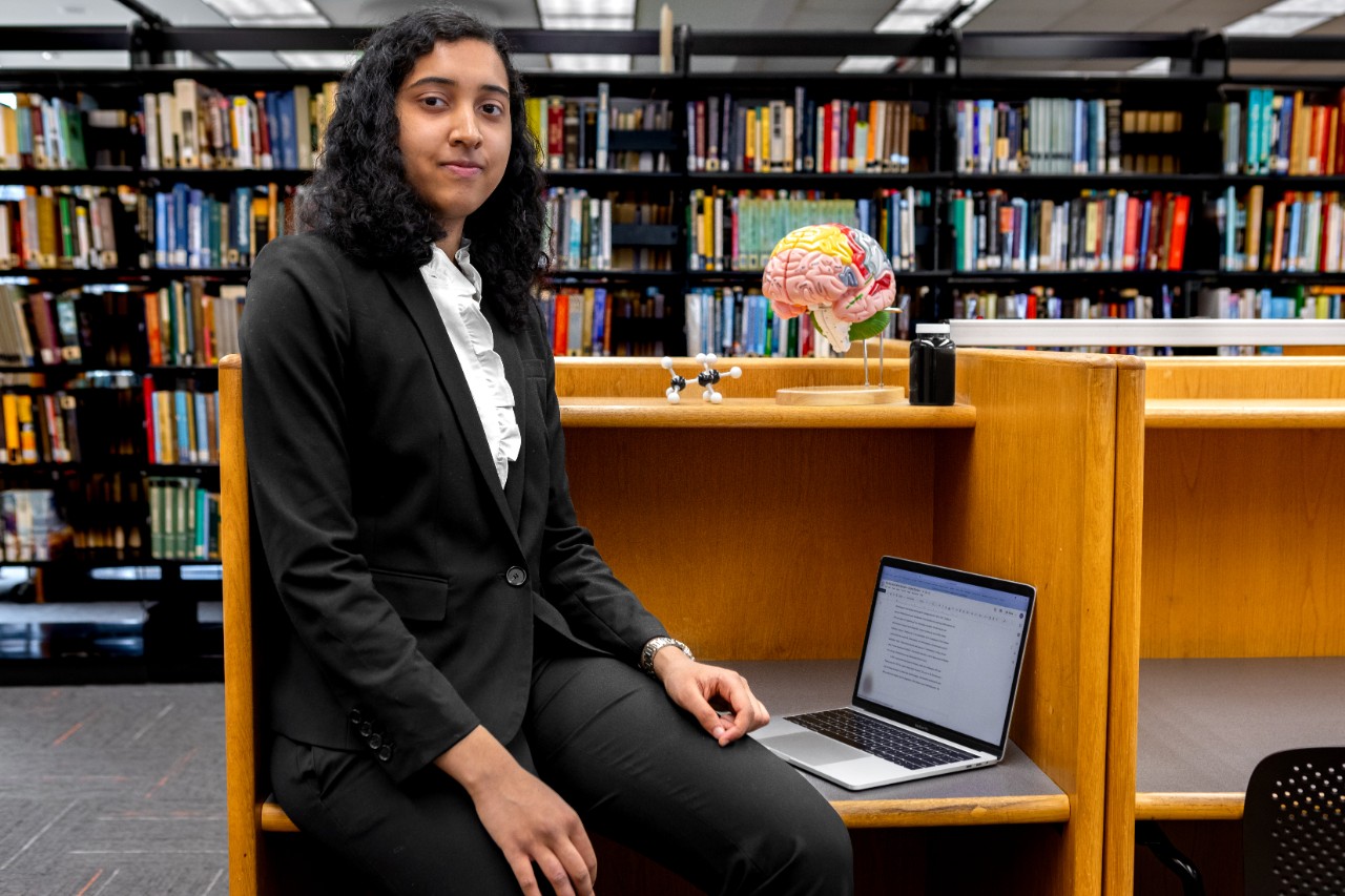 Portrait of Andrea Durham seated at a library desk. A laptop and a brain model are on the desk, with rows of bookshelves behind her.