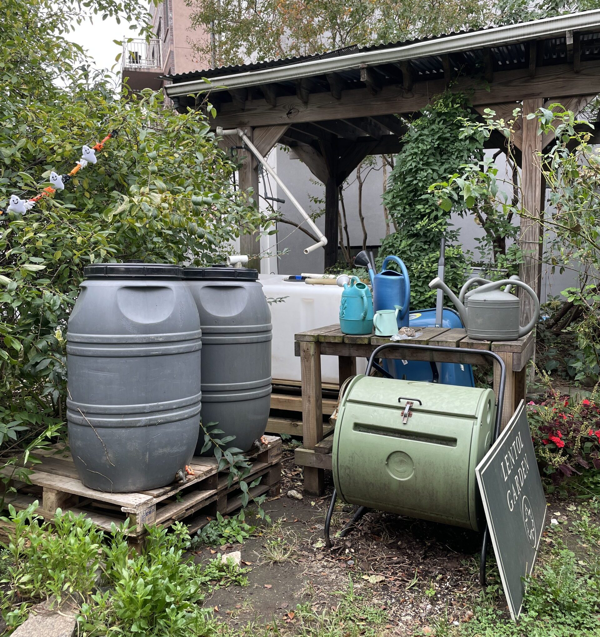 A small garden area with various gardening tools and equipment, including large gray rain barrels, watering cans, and a green compost bin on a stand. A wooden shelter is in the background, and a sign that reads 