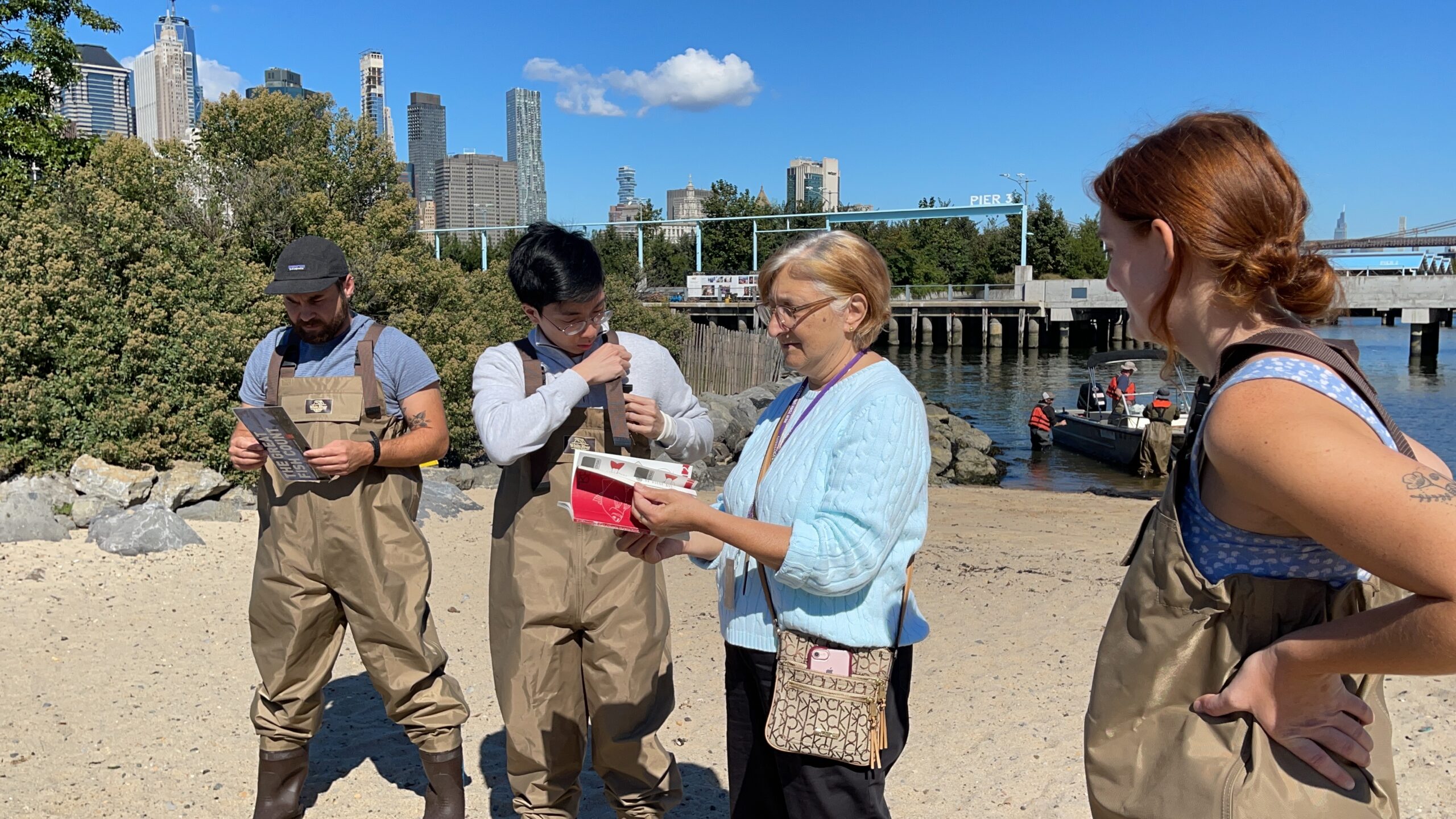 A group of people wearing waders are standing on a shore near a body of water. One person is holding a small red book, while others appear to be focused on their gear.