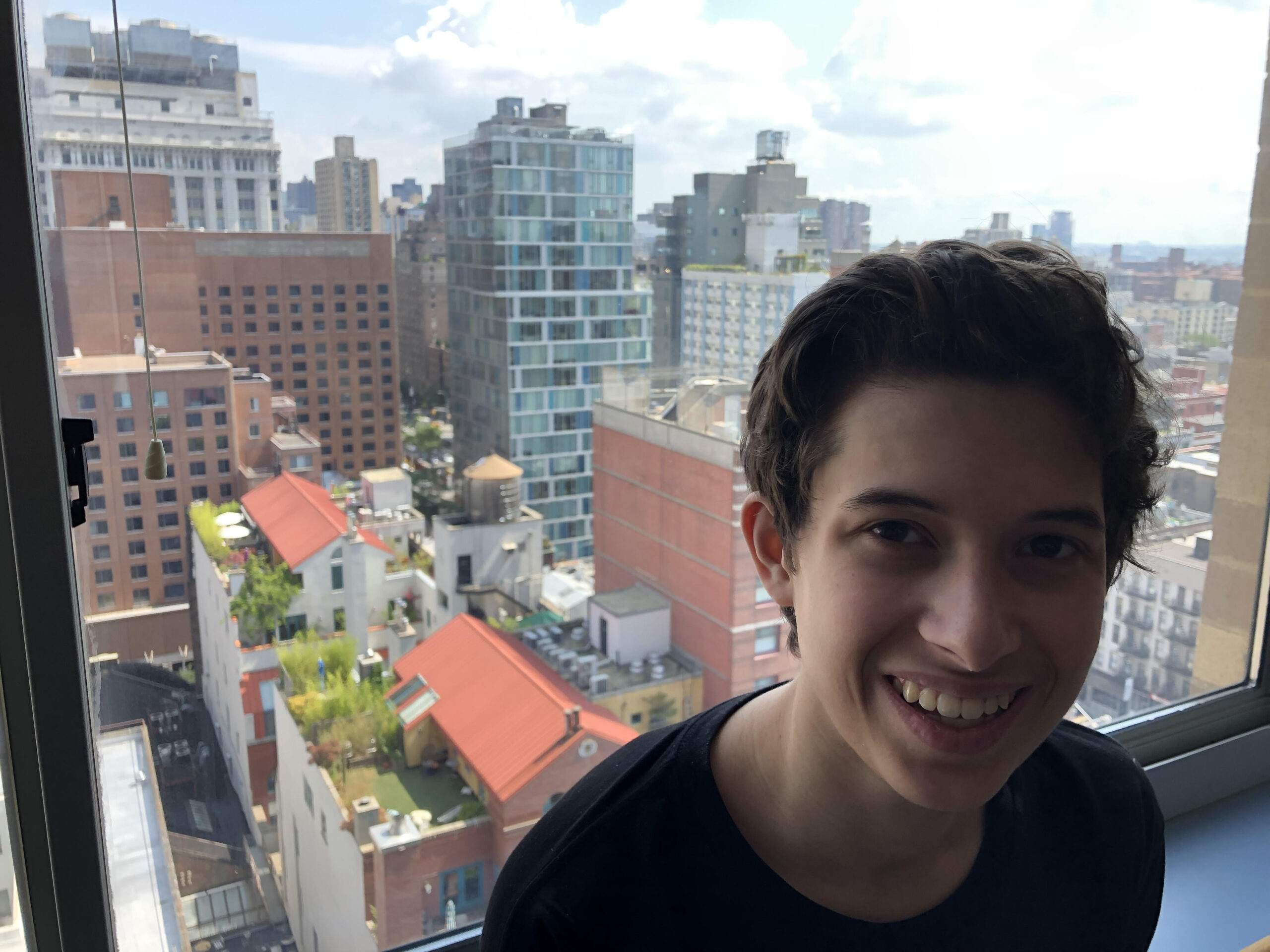 A teenager smiles in front of a window overlooking New York City after Tisch Summer High School Program move-in.